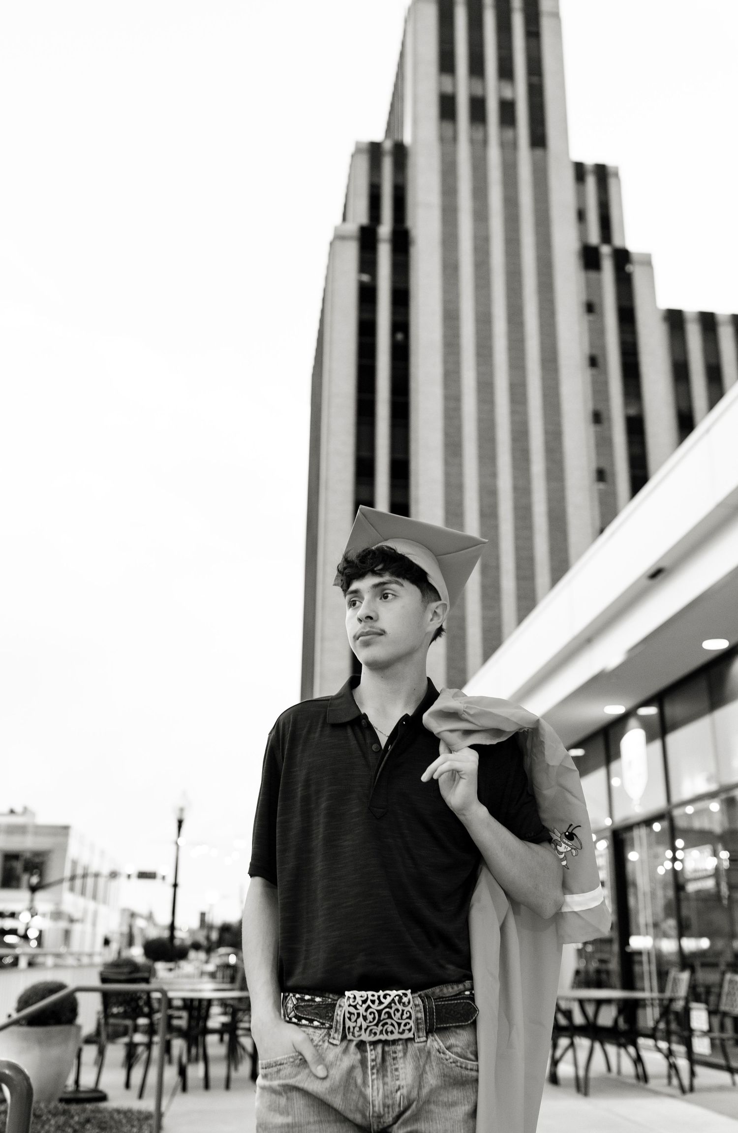 Black and white photo of male graduate holding cap near tall urban building downtown.