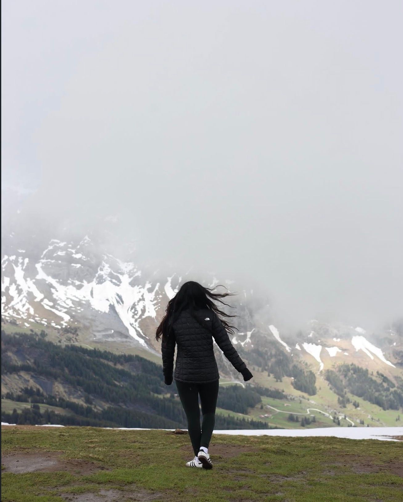 Woman walking on mountain plateau with snow-capped peaks and misty fog in background.
