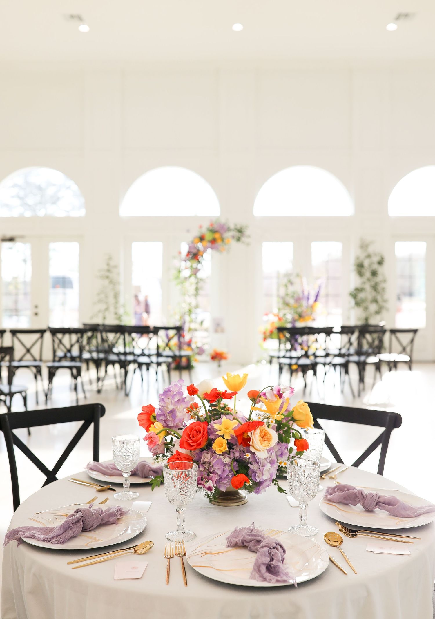 Elegant wedding reception hall with colorful floral centerpieces, lavender napkins, and gold cutlery on white tables.