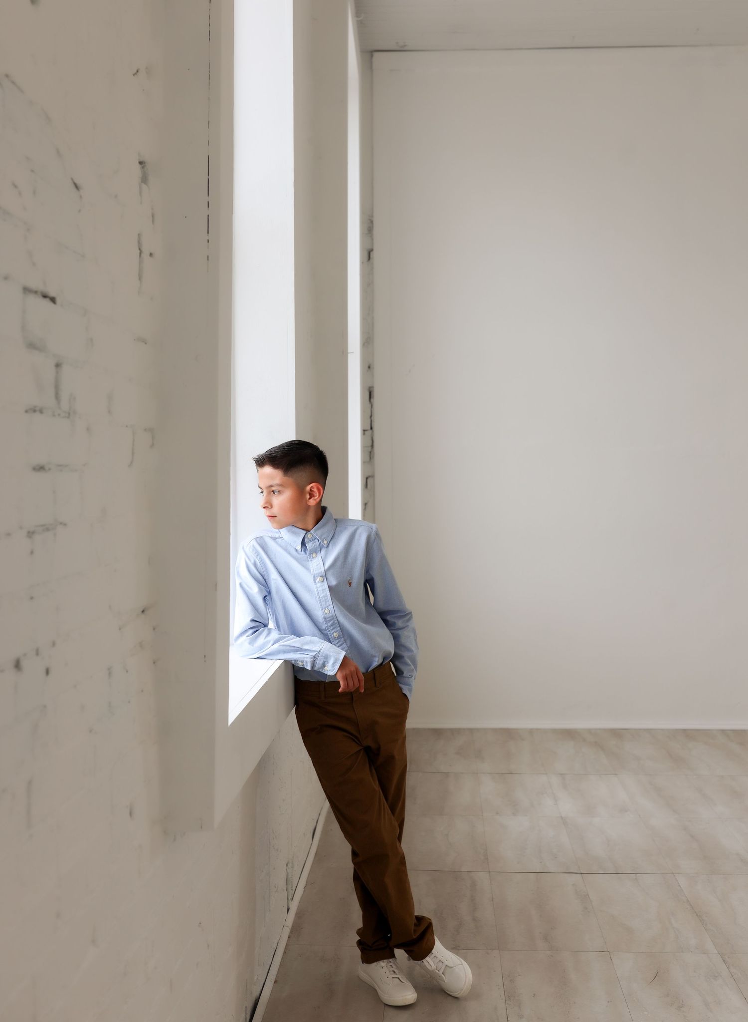 Teenage boy in light blue shirt and brown pants leaning against white wall beside tall narrow window.