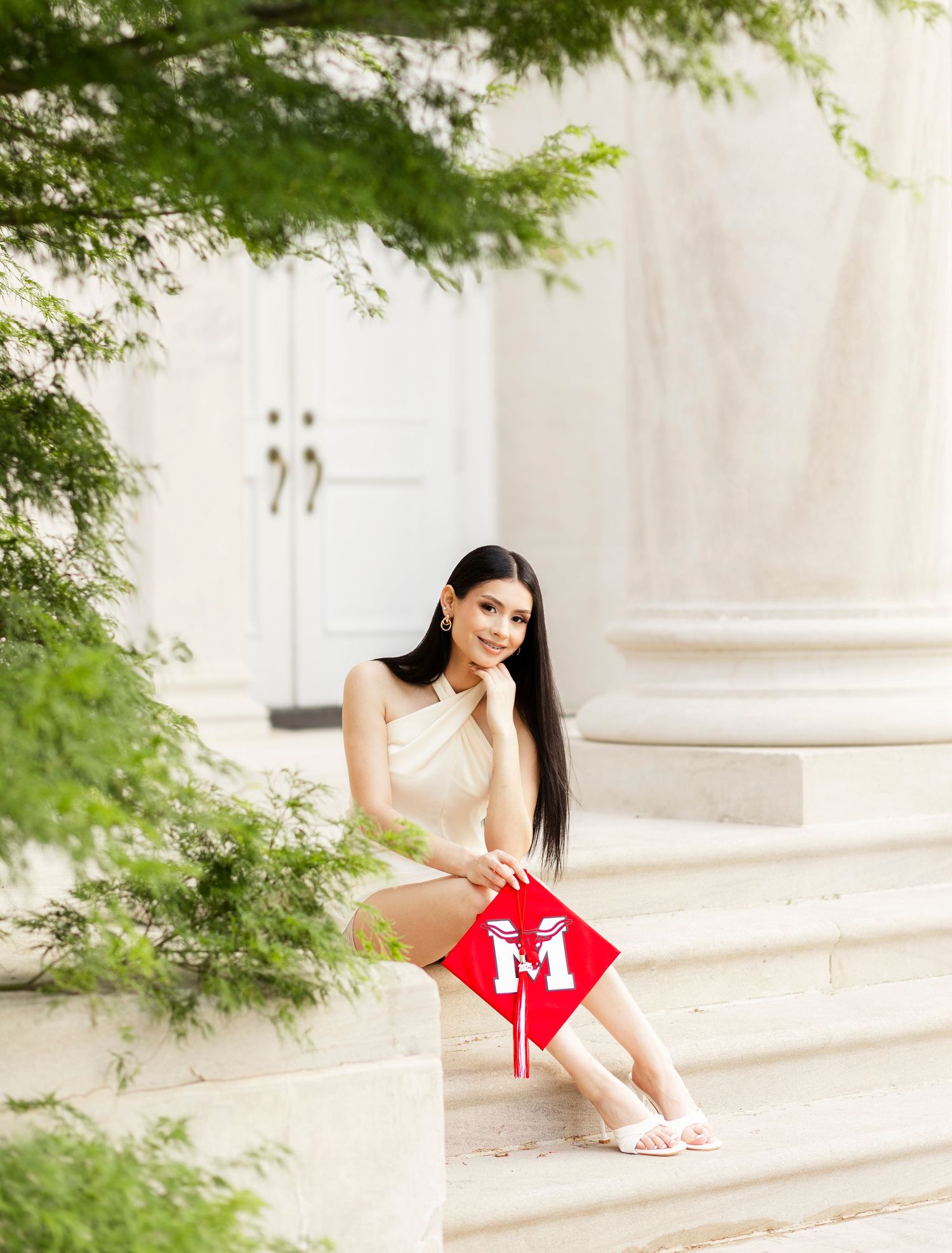 Woman in cream dress sits on marble steps holding red graduation cap beside lush greenery.