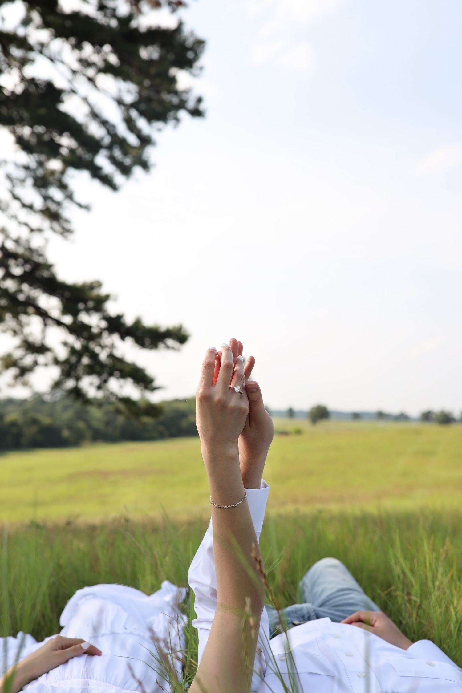 Couple lying in grass holding hands raised up with engagement ring visible against open field sky.