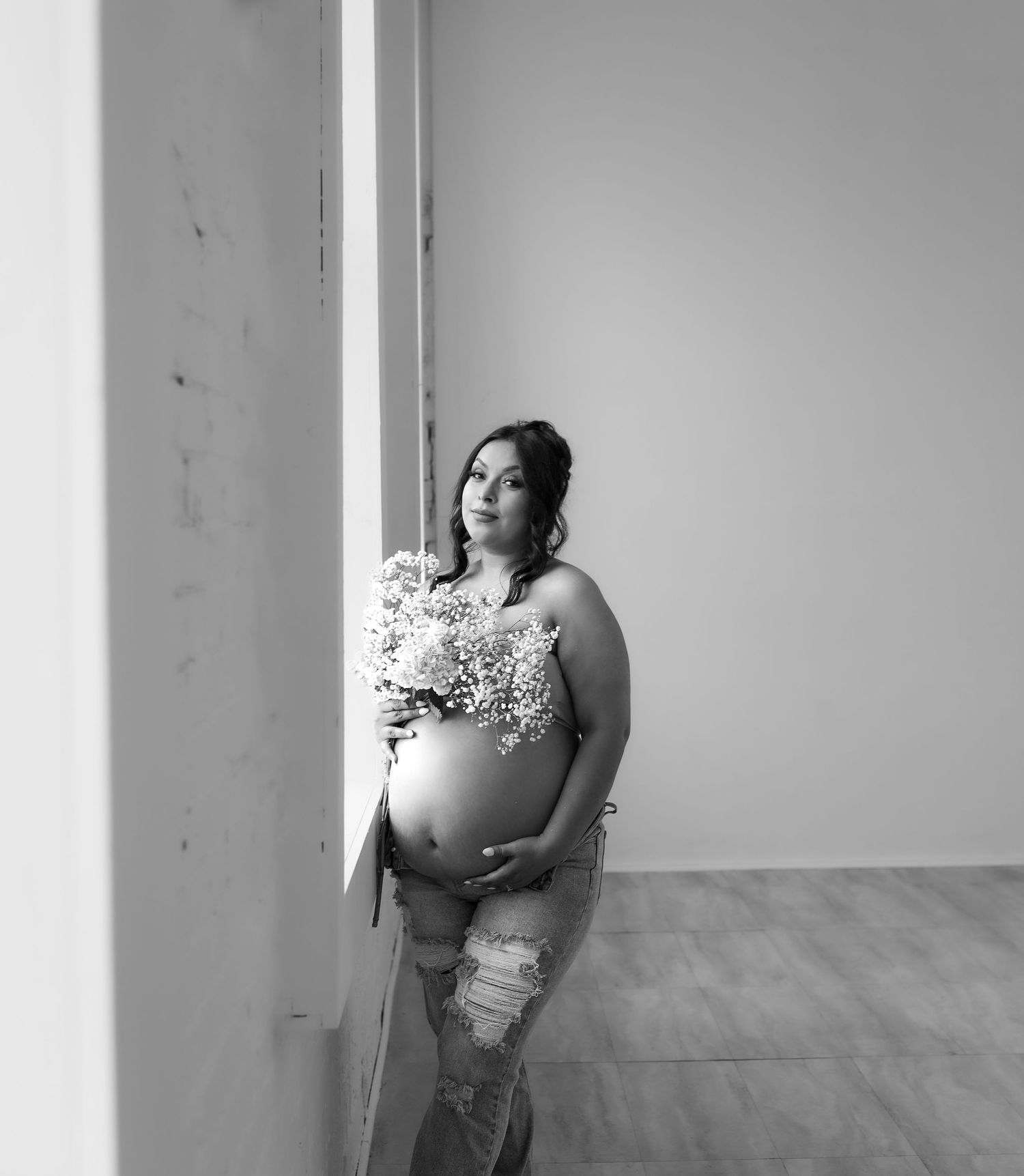 Pregnant woman holding white flowers against wall, black and white maternity portrait in studio.