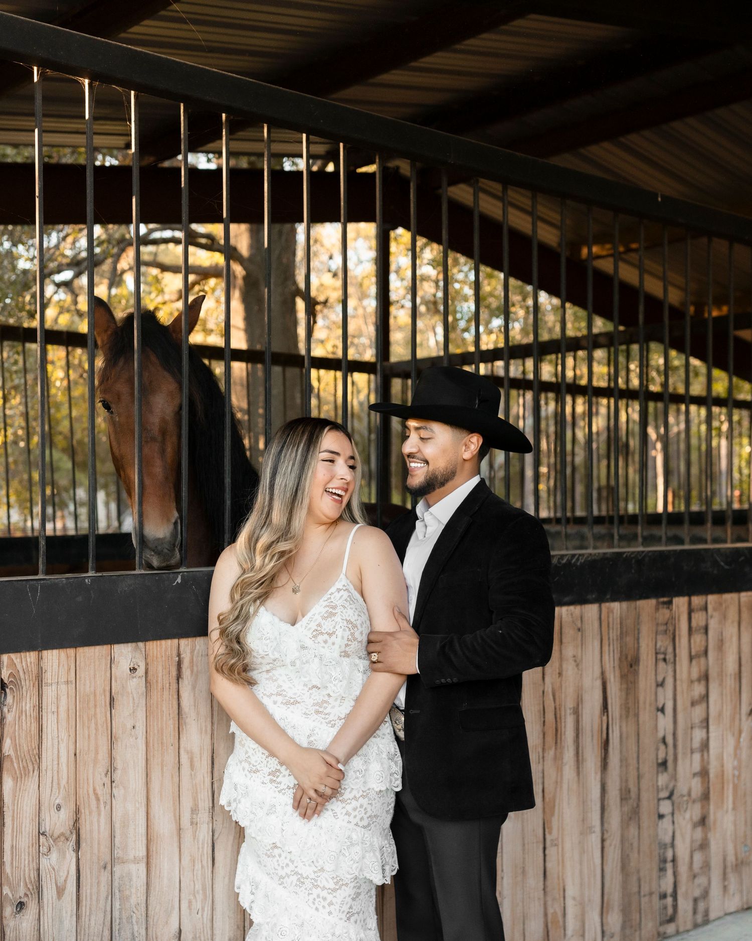 A smiling couple stands together in front of a horse stable, woman in white lace dress, man in black cowboy hat.