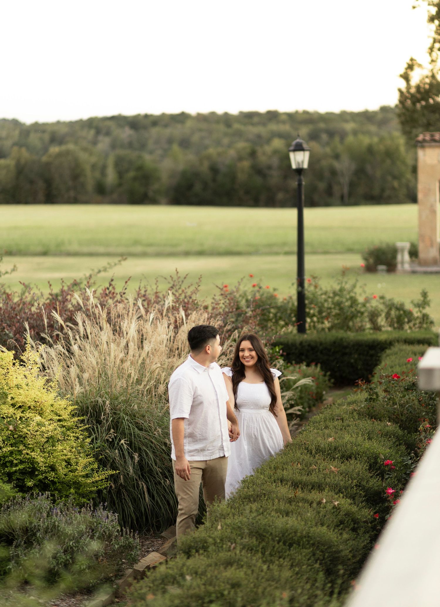 Couple walking hand in hand through lush garden path with green fields and trees in background.