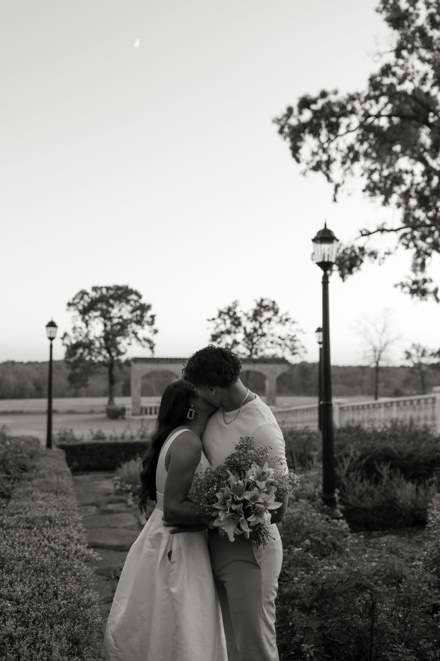 Black and white photo of couple kissing in garden with bouquet at dusk.