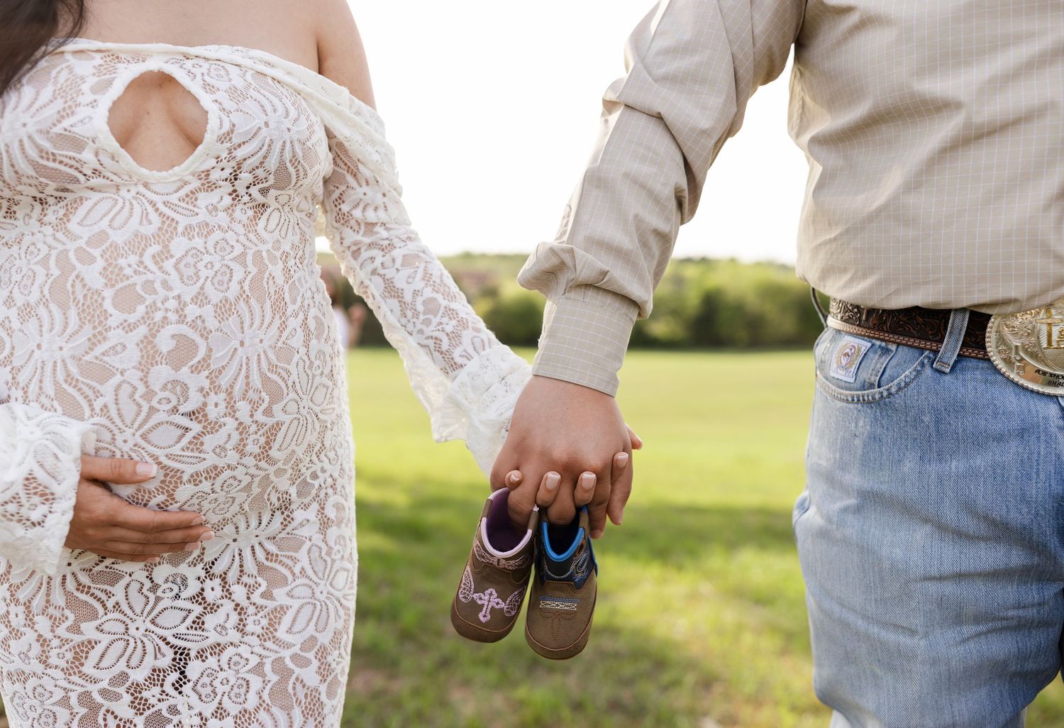 Couple holding tiny baby boots while standing in green field, woman in white lace maternity dress.
