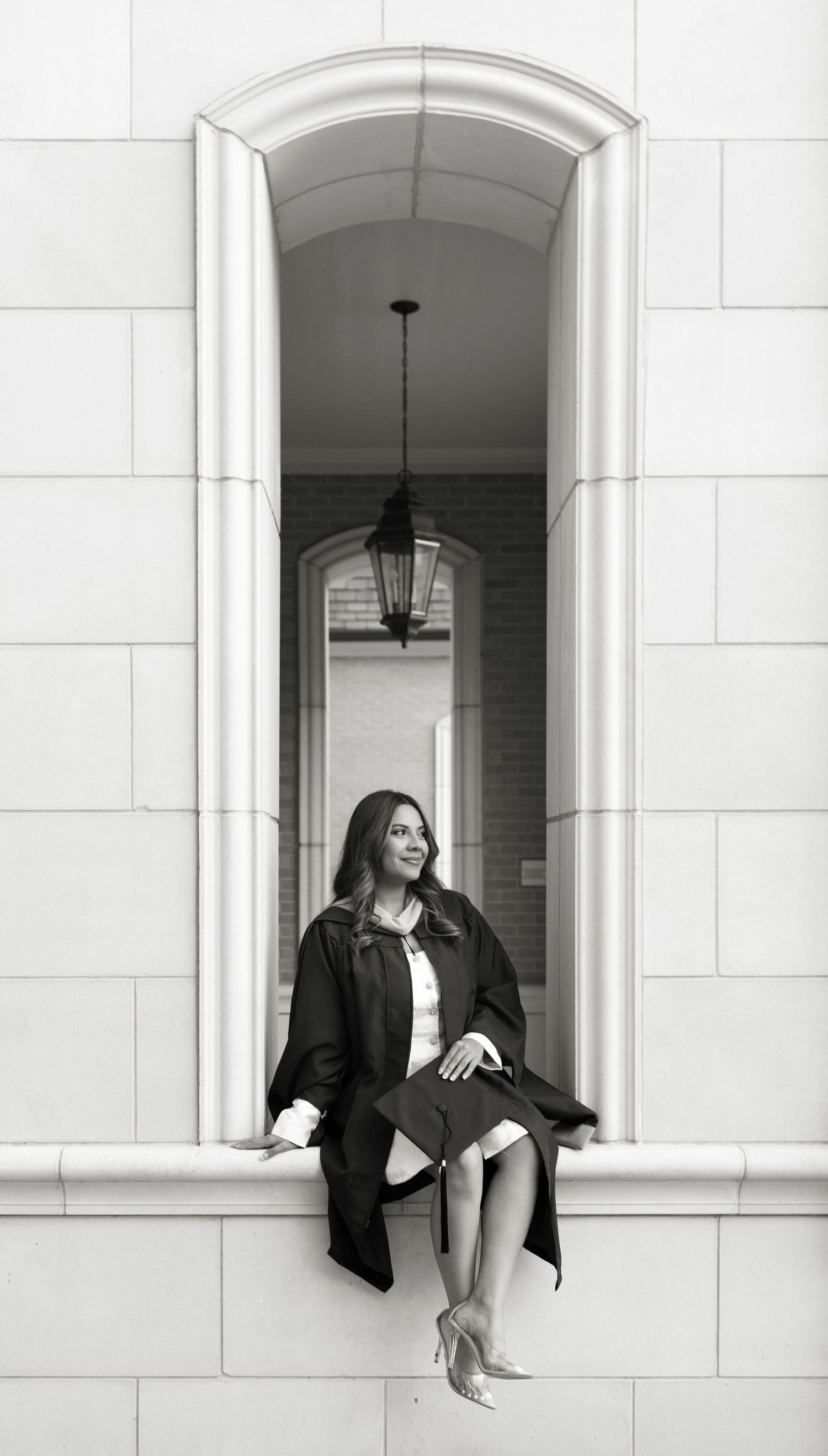 Black and white photo of graduate in gown sitting in arched window niche of white stone building.