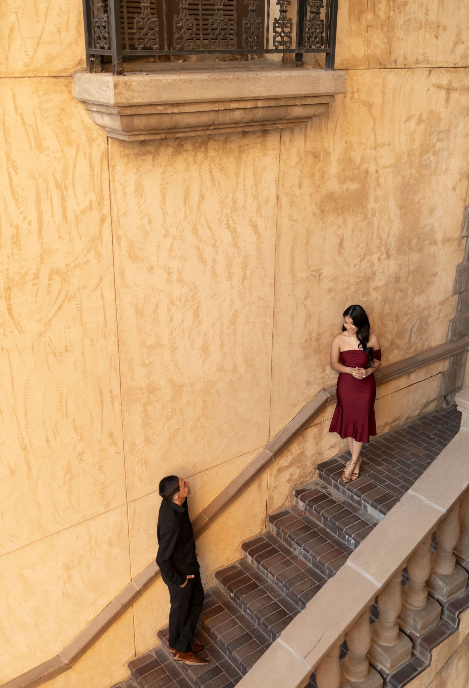 Couple on ornate staircase, woman in burgundy dress descending toward man in black.