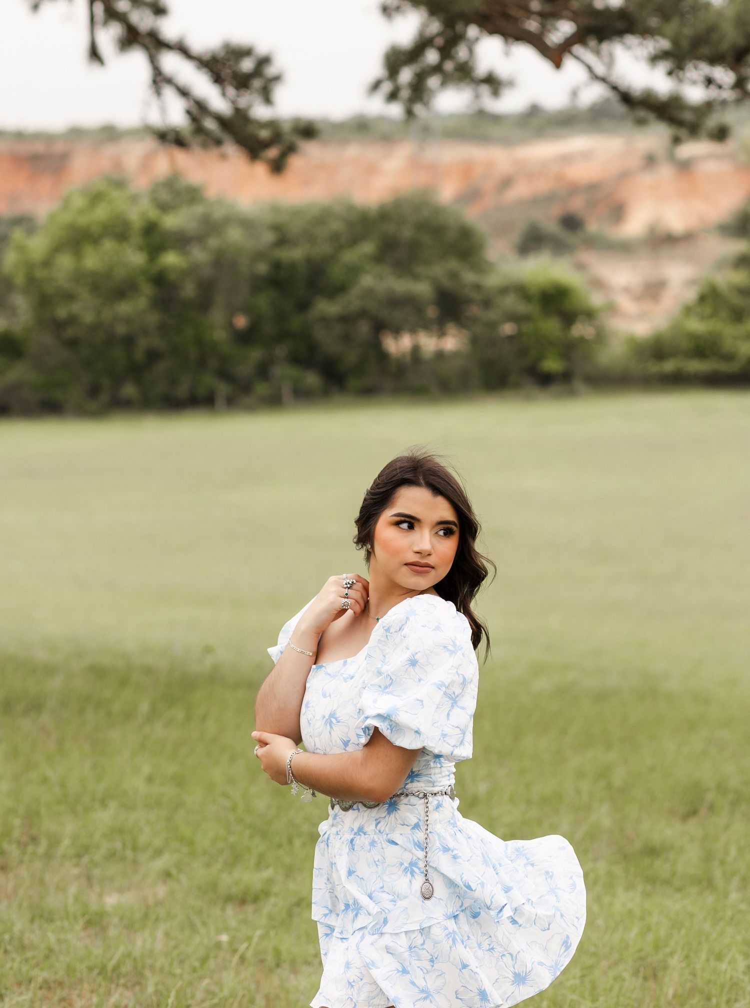Young woman in blue floral dress poses in green field with red clay cliffs in background.
