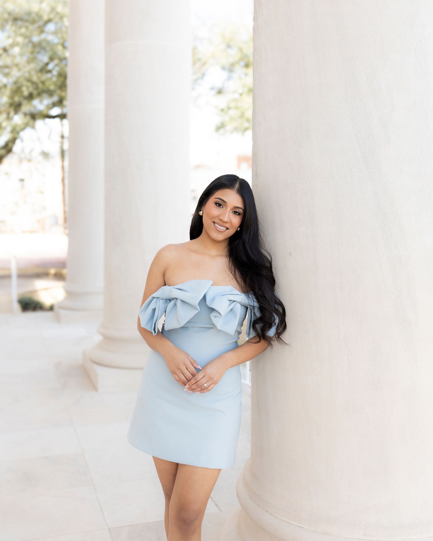 Young woman with long dark hair in light blue ruffled off-shoulder mini dress leans against white column.
