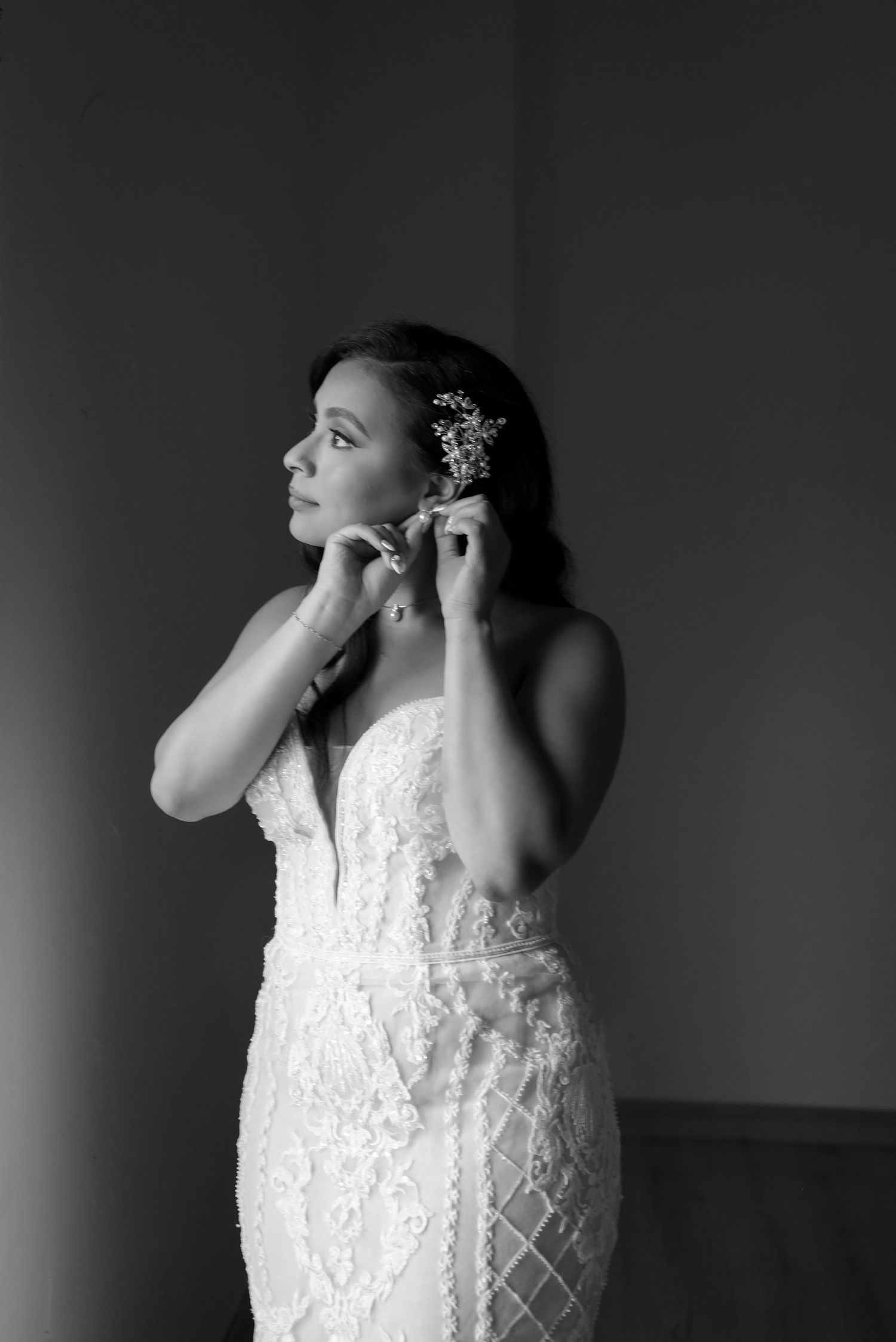 Black and white portrait of bride in lace gown putting on earrings with floral hair accessory.