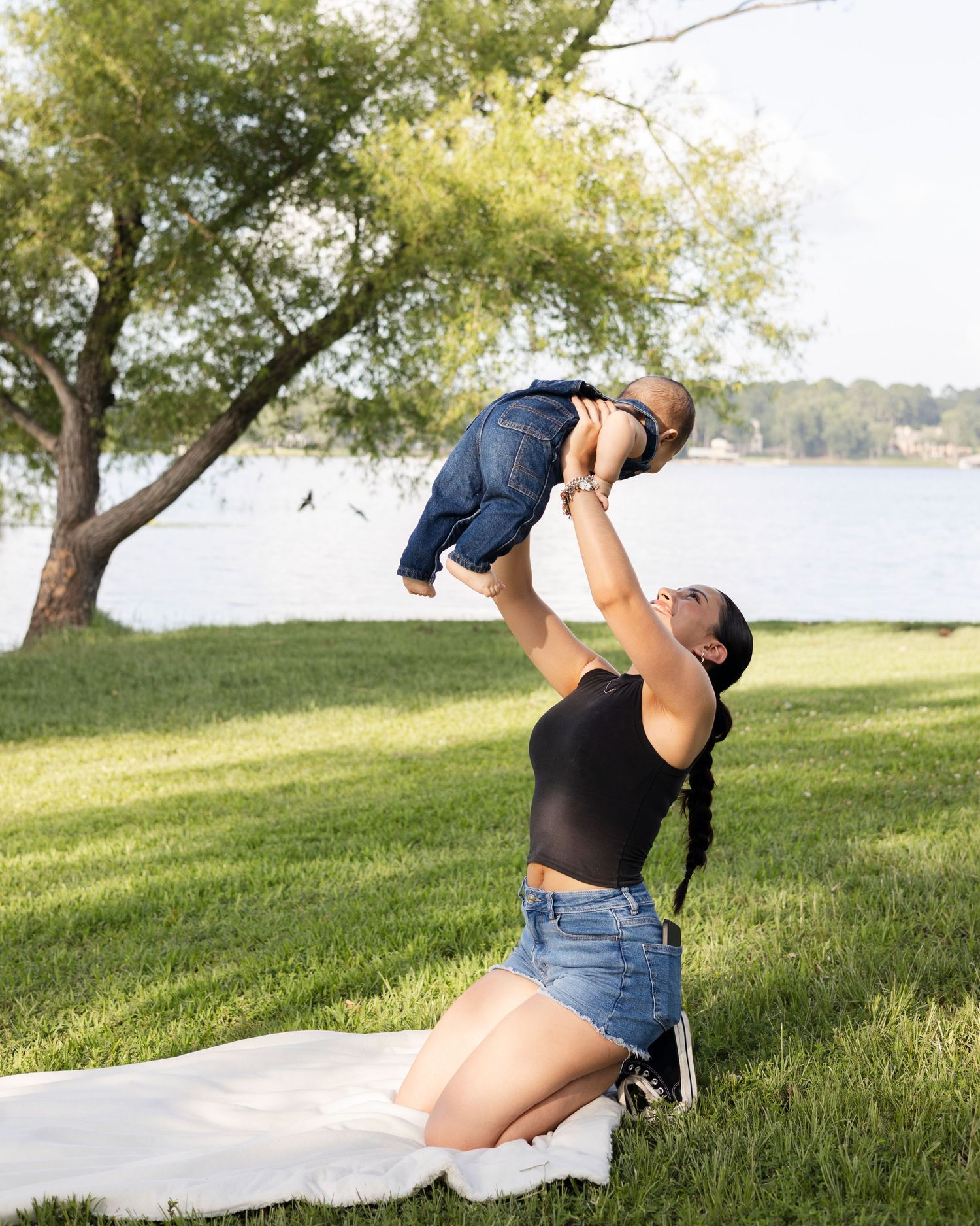 Mother kneeling on grass lifting her baby boy in the air near a lakeside park.