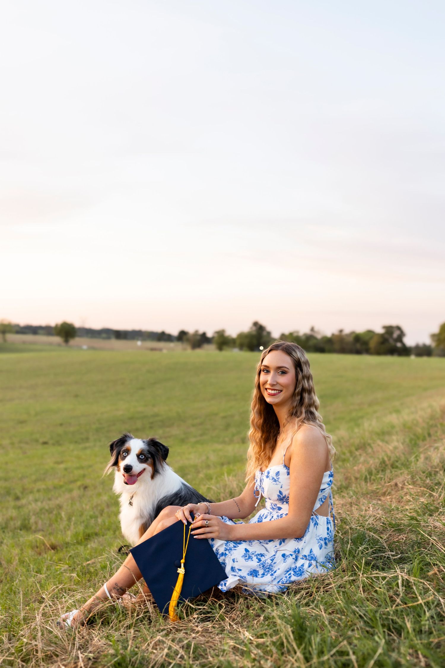 Young woman in floral dress sits with her Australian Shepherd dog wearing a graduation cap in a green field at sunset.