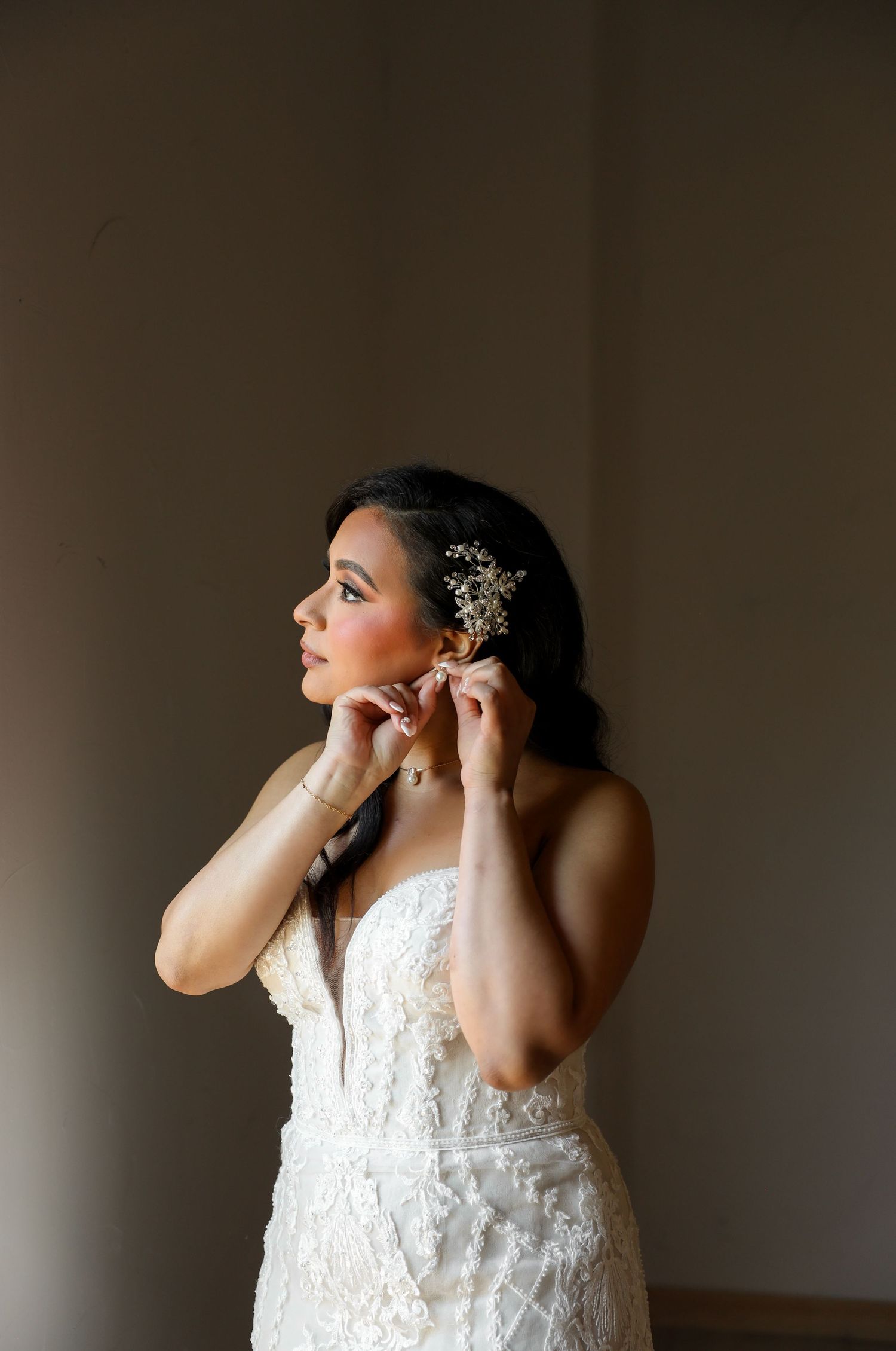 Bride in strapless lace wedding gown putting on earrings with baby breath hair accessory in soft light.