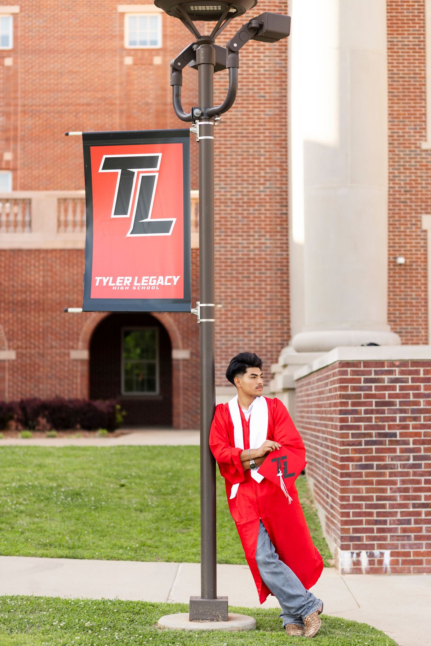 Male graduate in red cap and gown leaning against Tyler Legacy High School banner pole.