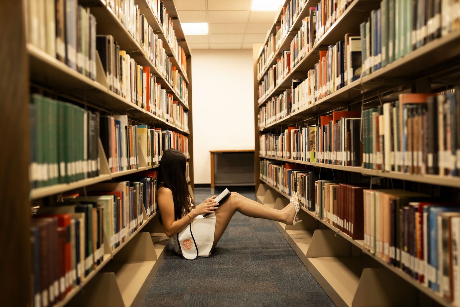 Student sitting on library floor between bookshelves reading a book in a college library.