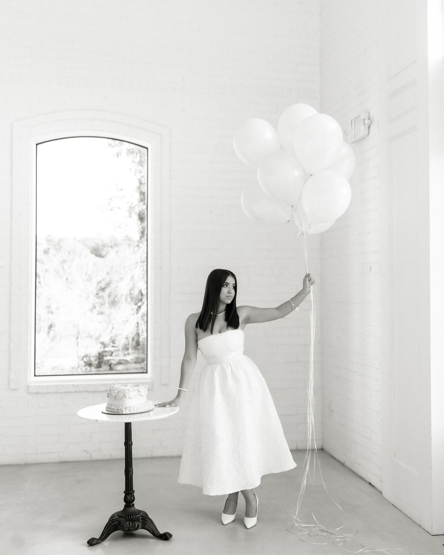 Black and white photo of woman in white midi dress holding balloons beside small cake table.