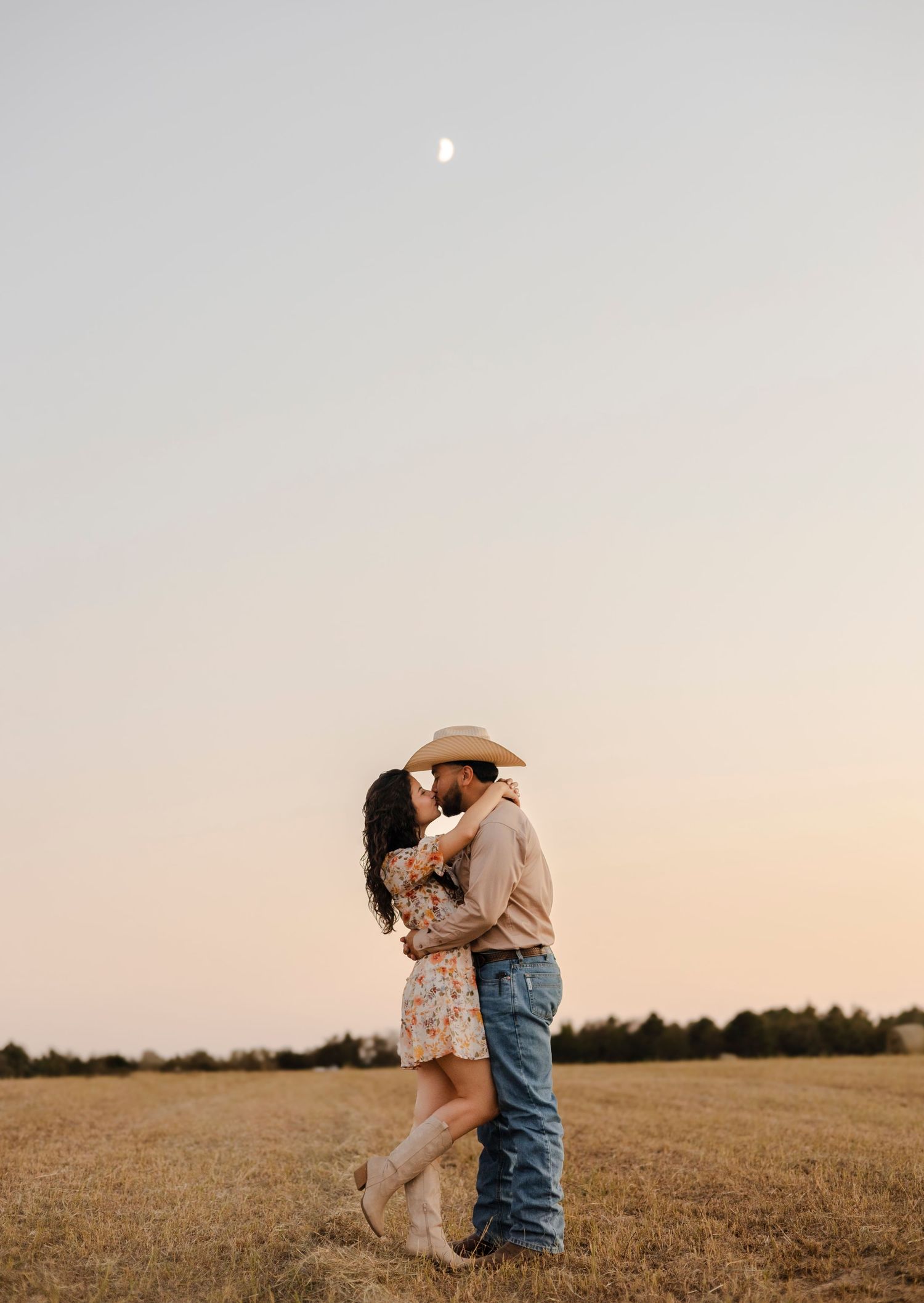 Couple sharing a romantic kiss in a golden field at dusk with a crescent moon above.