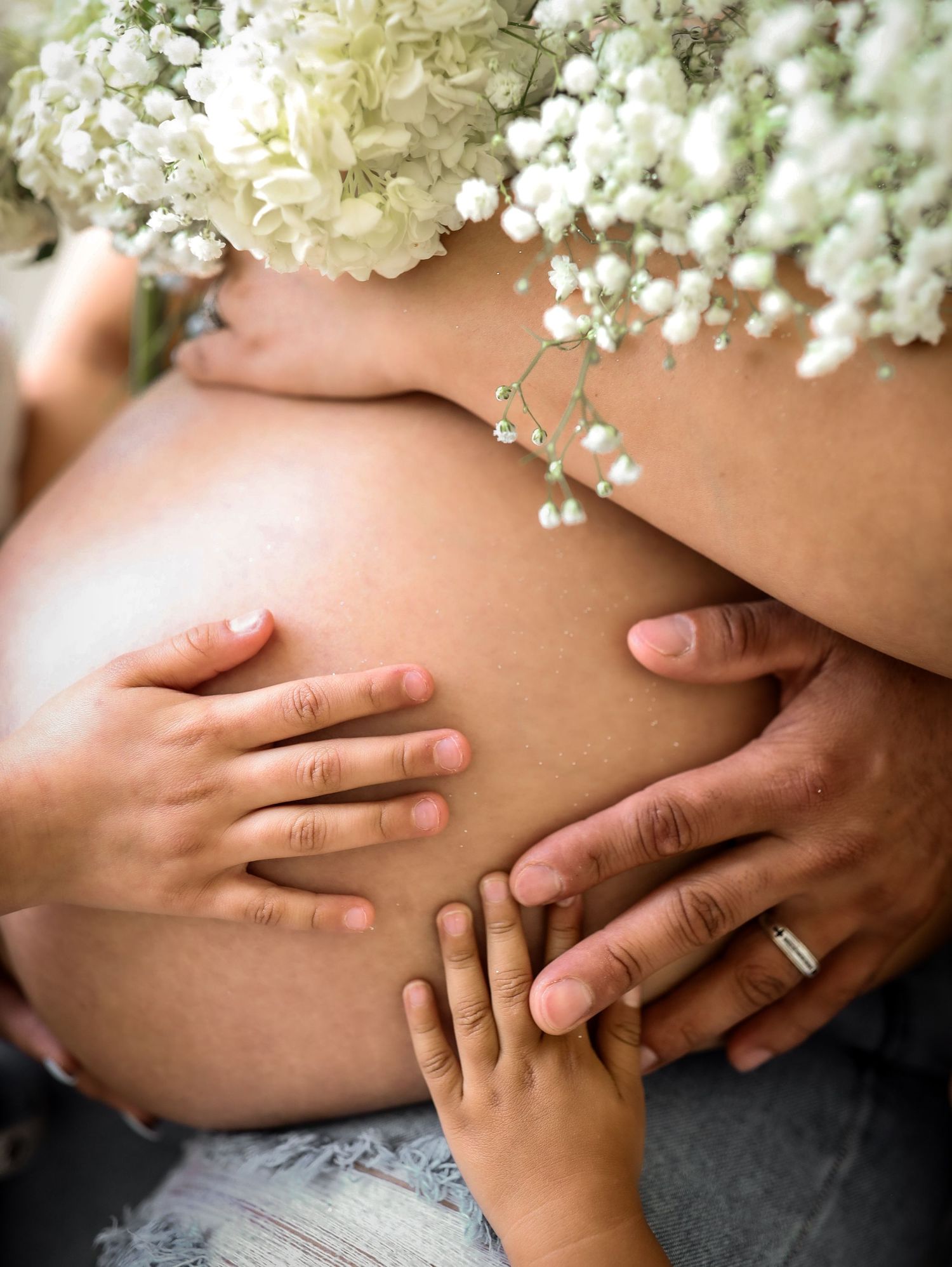 Close-up of pregnant belly with family hands and white floral bouquet arrangement.