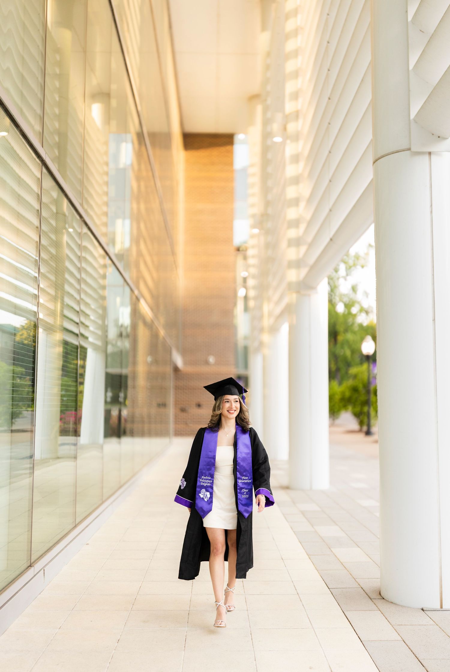 Female graduate in black cap and gown with purple stole walks confidently along modern university building colonnade.