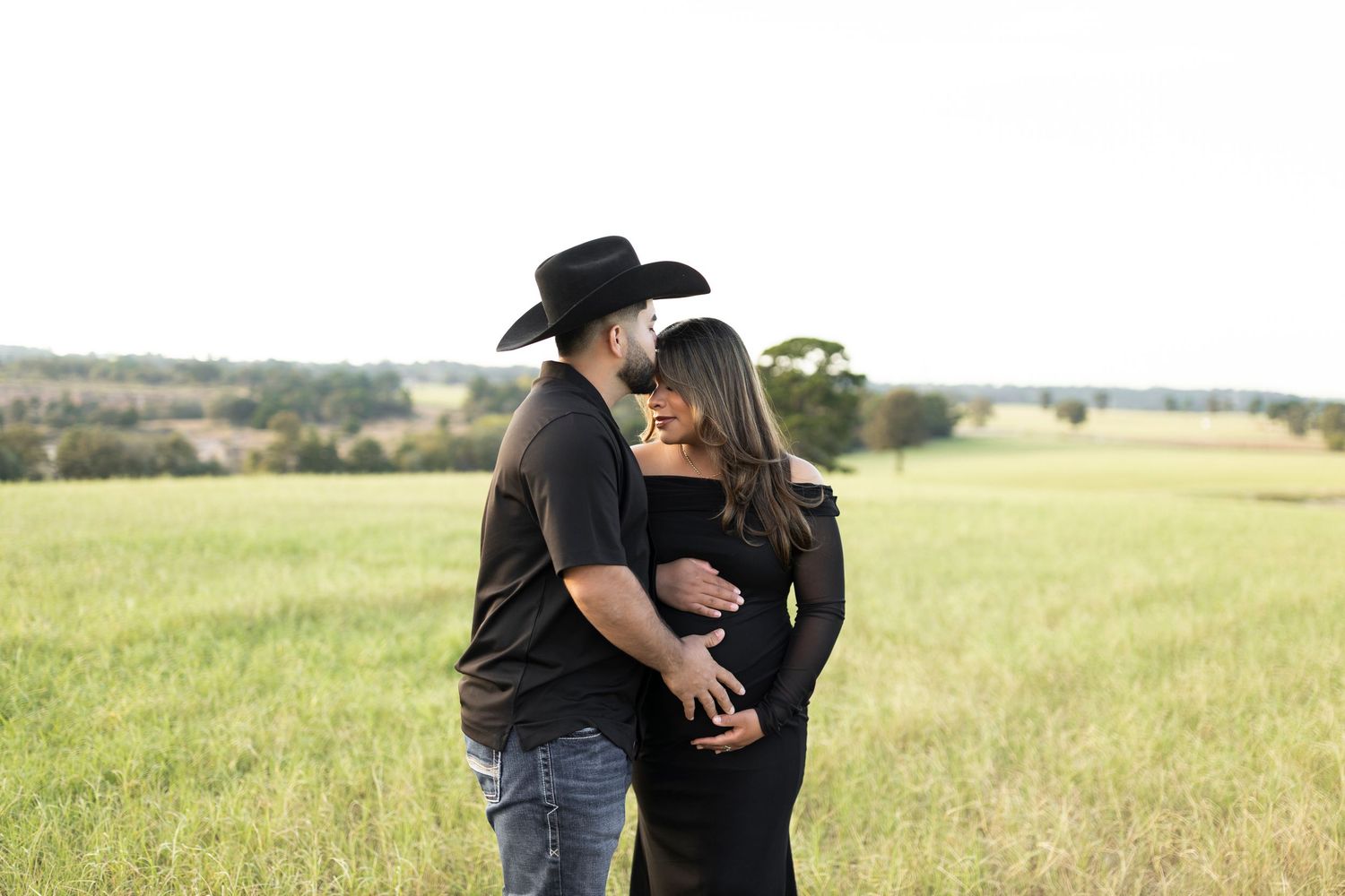 Couple embracing in a green field, man in black cowboy hat holding pregnant partners belly.