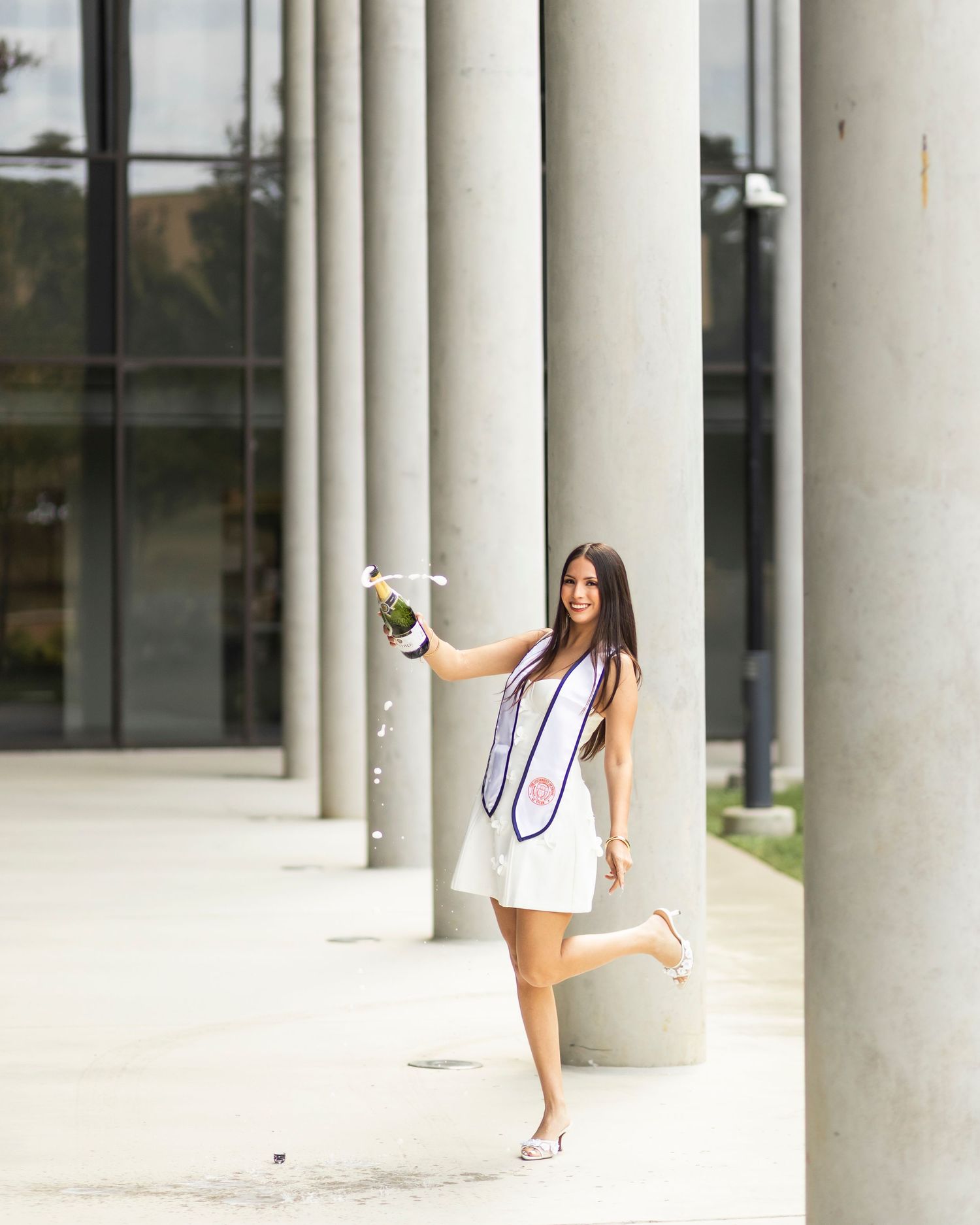 Female graduate in white dress pops champagne bottle while posing between concrete columns outdoors.