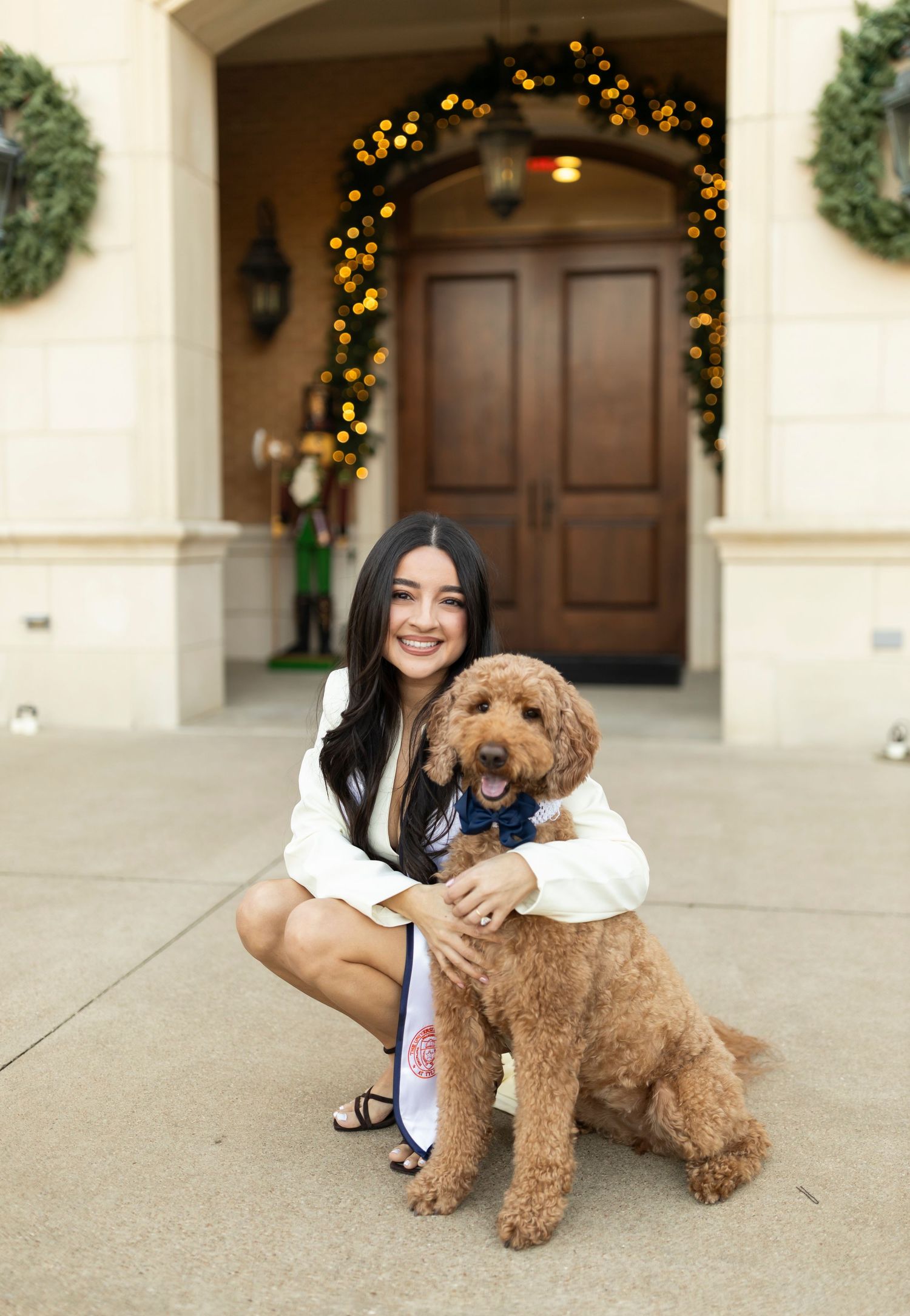 Smiling female graduate in white outfit crouches with golden doodle dog outside decorated entrance.