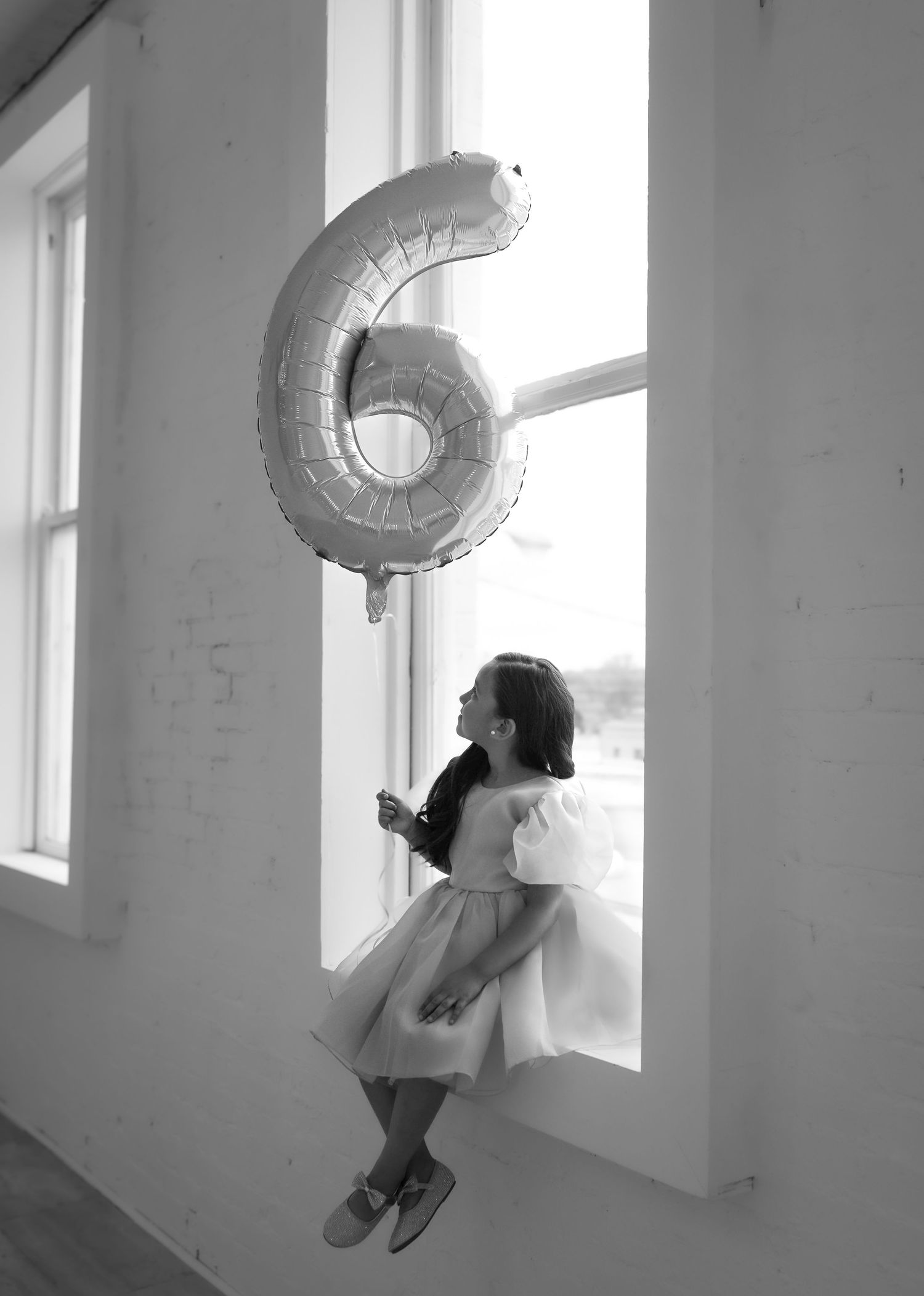 Black and white photo of young girl in tulle dress sitting on windowsill holding number 6 balloon.
