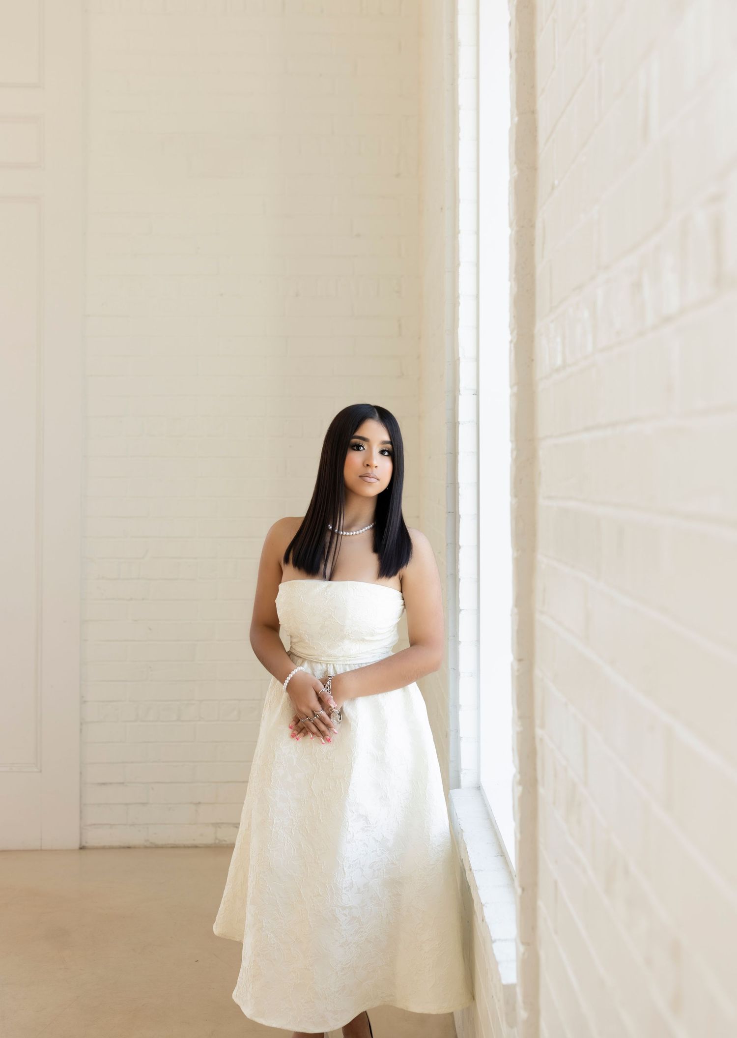 Elegant brunette woman in strapless white midi dress leaning against bright window on white brick wall.