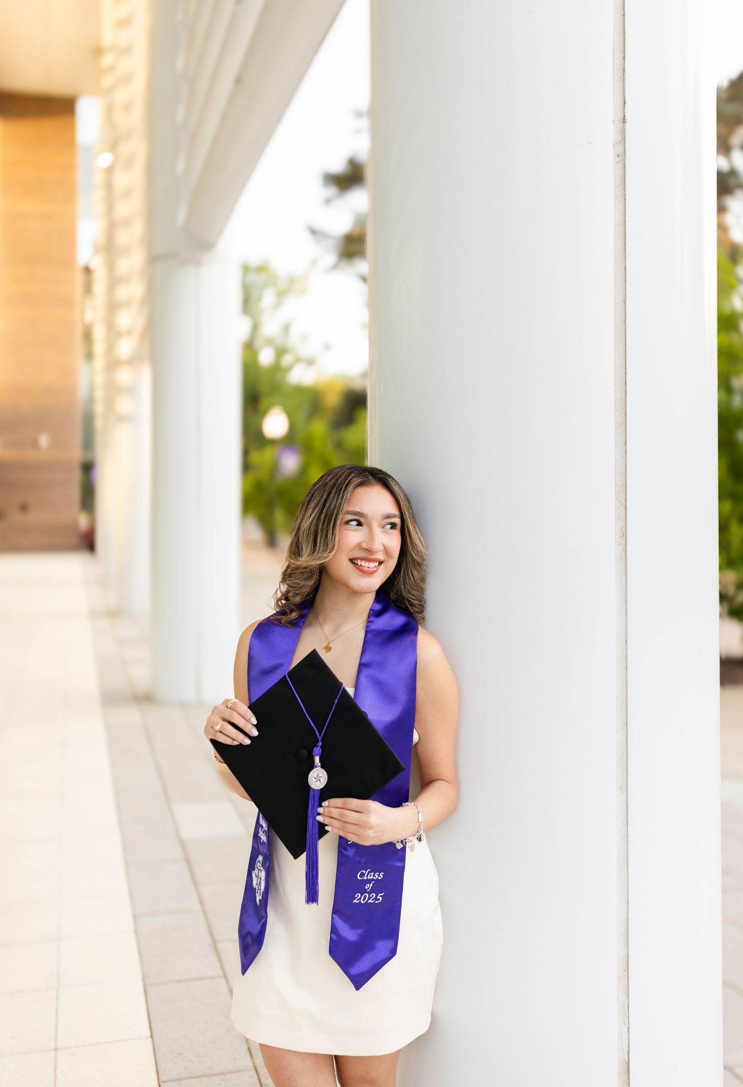 Smiling graduate leans against white column wearing purple Class of 2023 stole holding diploma.