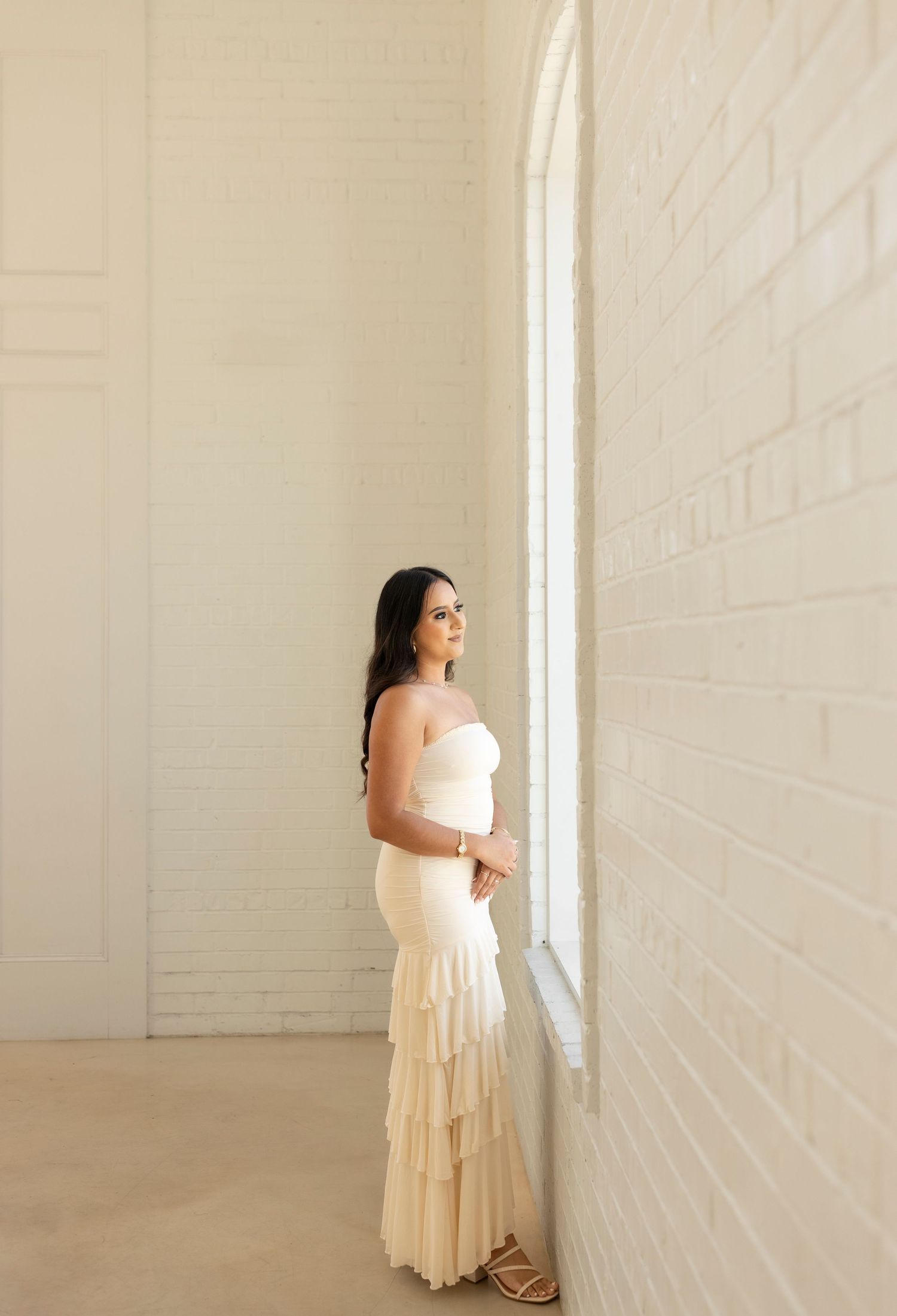 Pregnant woman in cream strapless ruffled gown stands by arched window in bright white brick room.