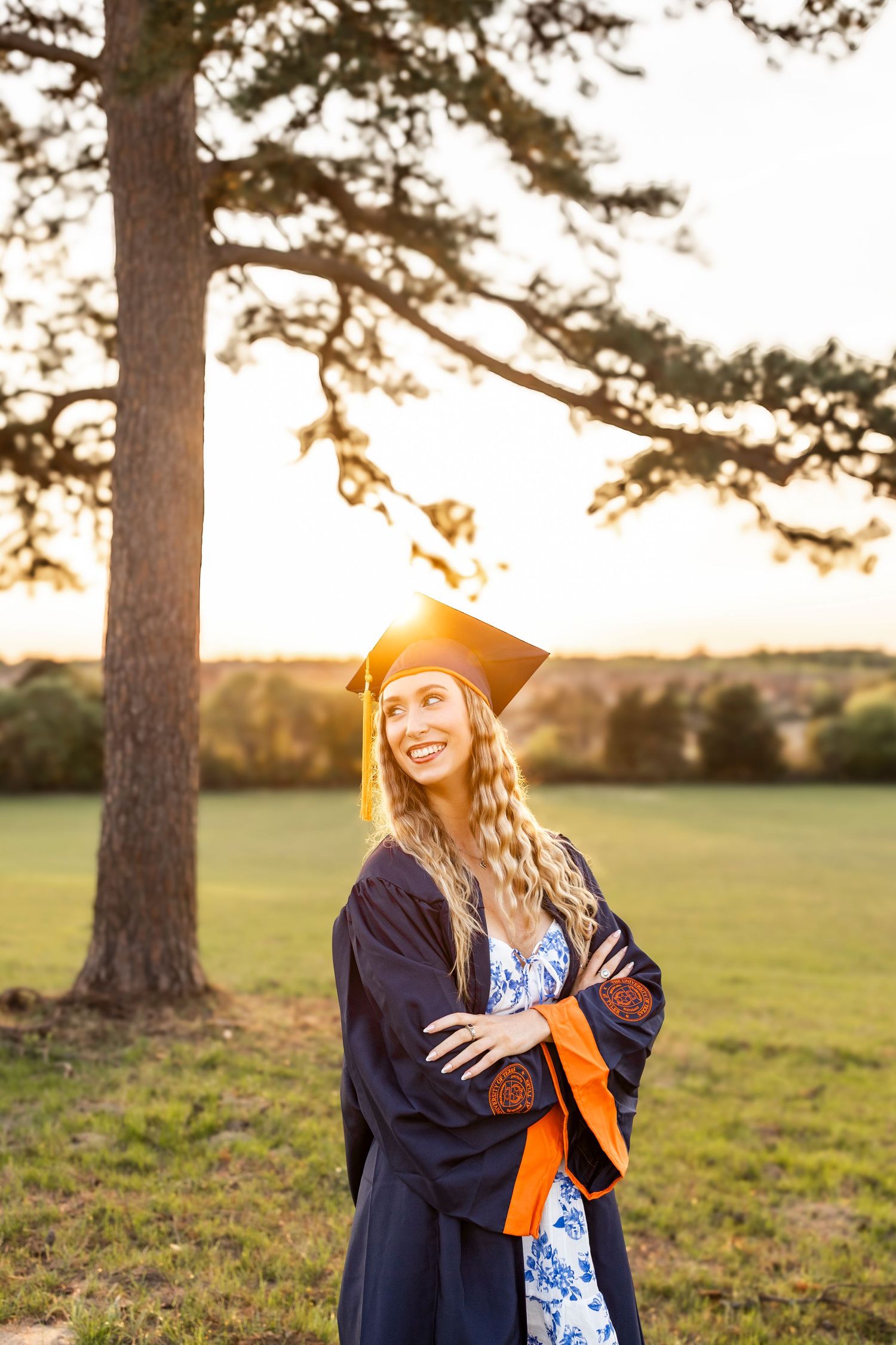 Smiling graduate in navy cap and gown with orange sash stands arms crossed in sunny park at golden hour.