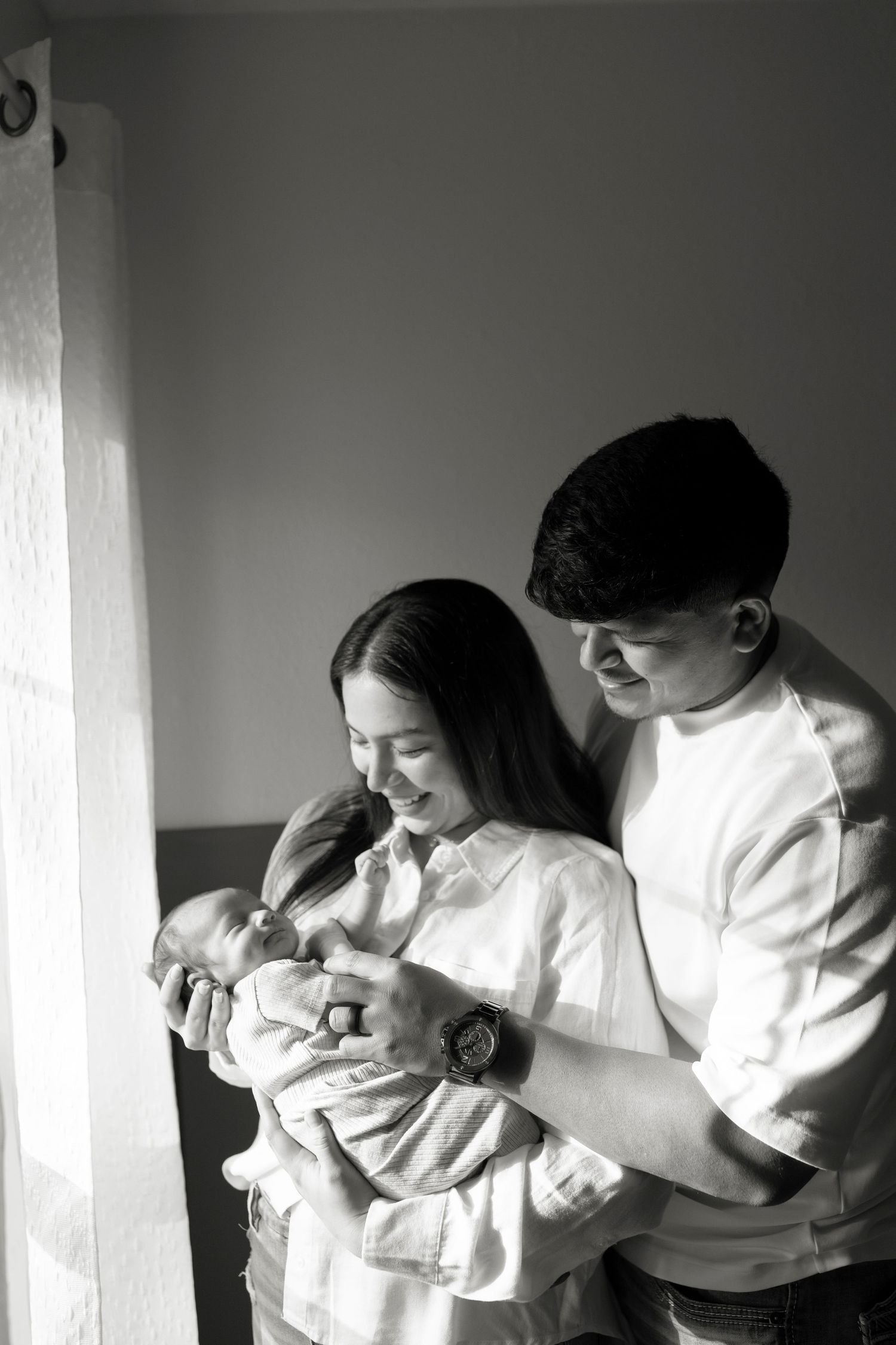 Black and white portrait of smiling new parents holding their newborn baby by a window.