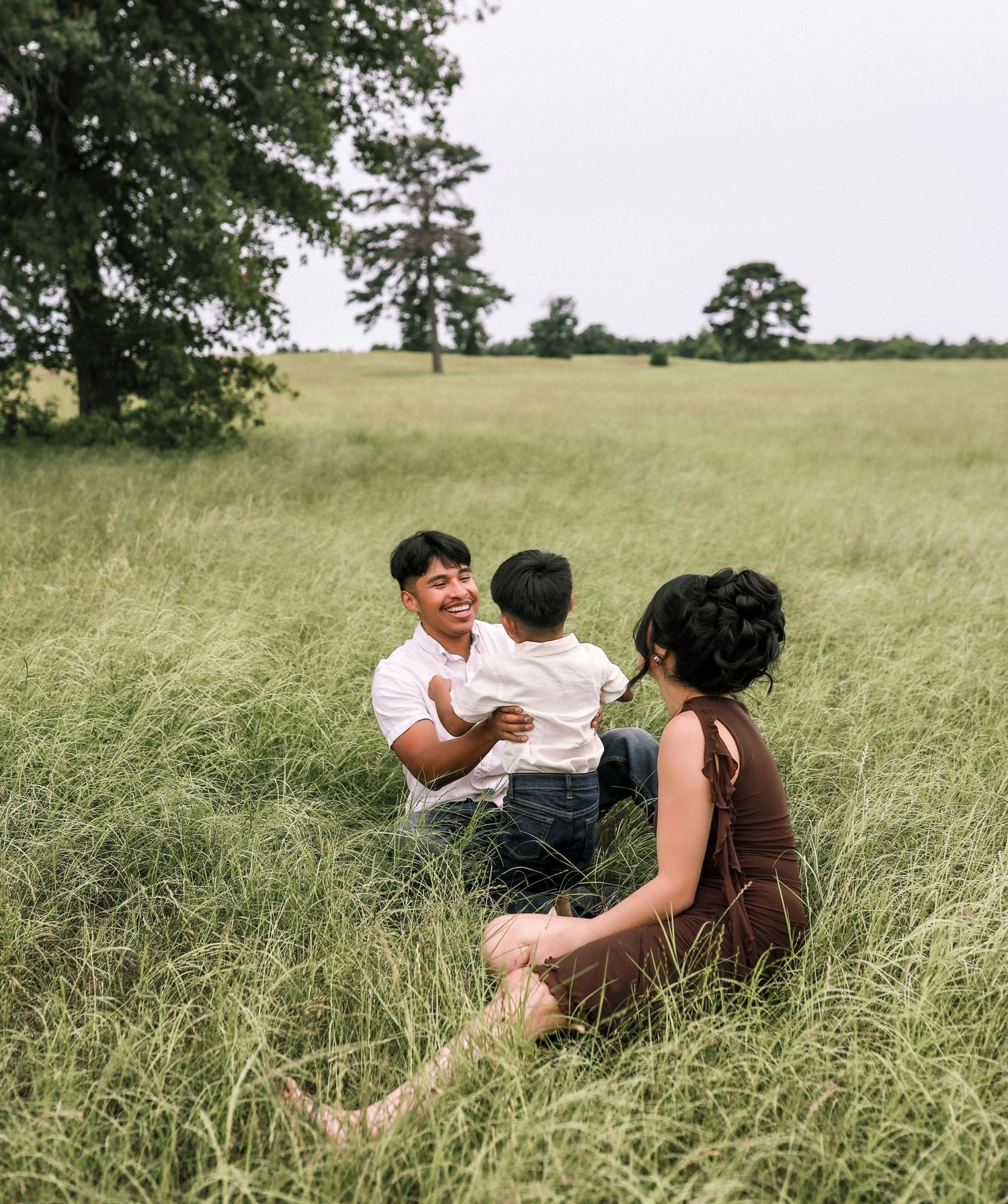 Happy family sitting in tall grass field, father lifting smiling toddler while mother watches lovingly nearby.