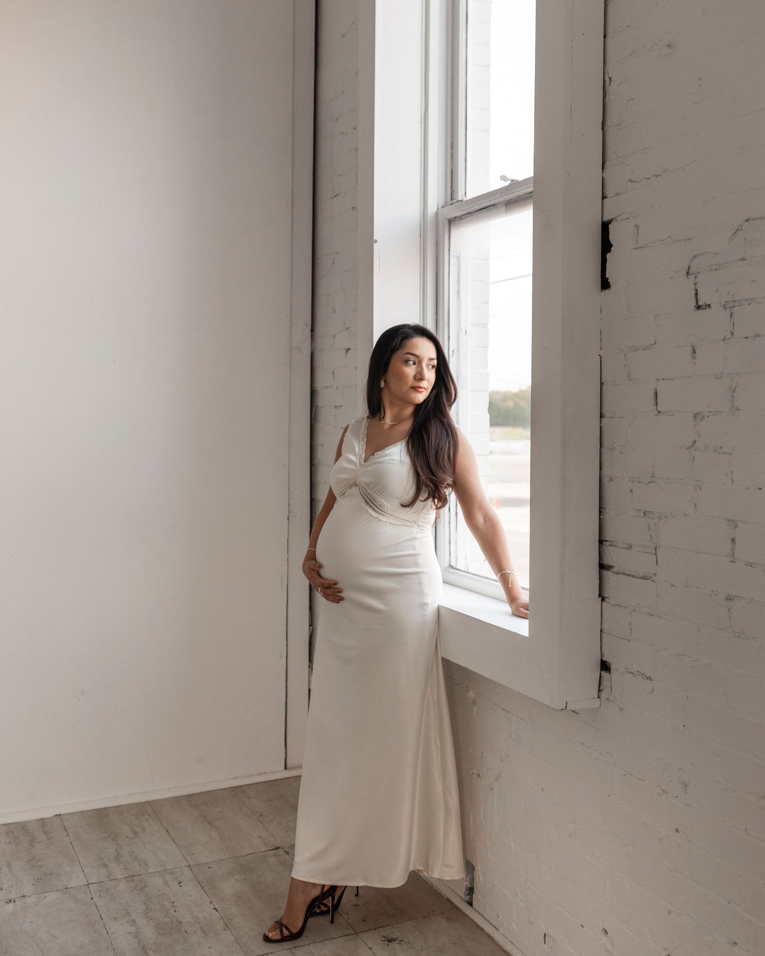 Pregnant woman in white satin gown posing by a bright window in a white brick studio.