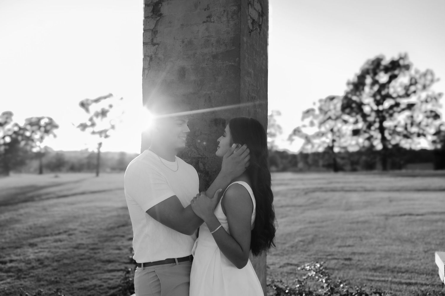 Black and white photo of couple sharing a kiss at golden hour beside a stone pillar outdoors.