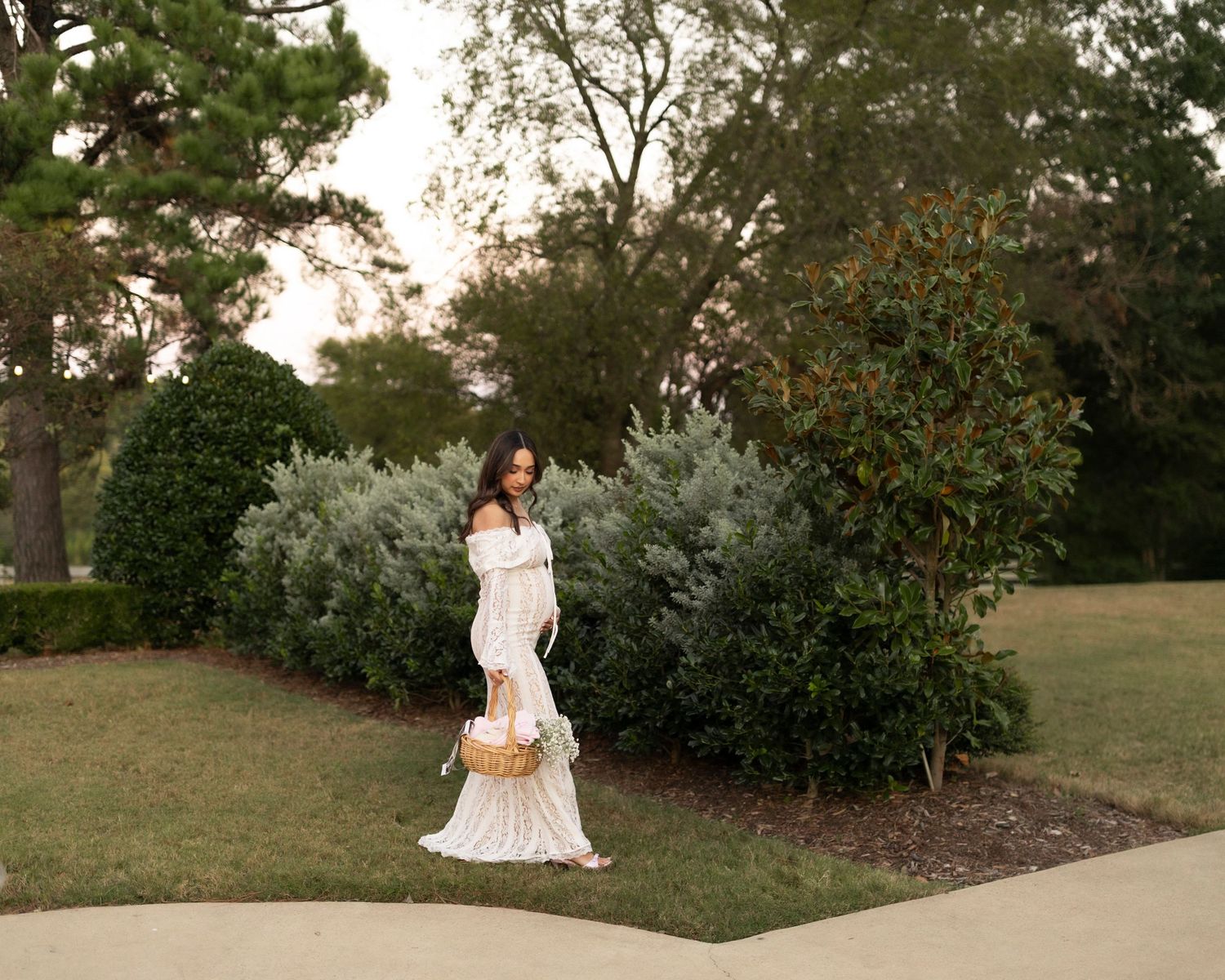 Pregnant woman in white off-shoulder gown walking through lush green garden at dusk.