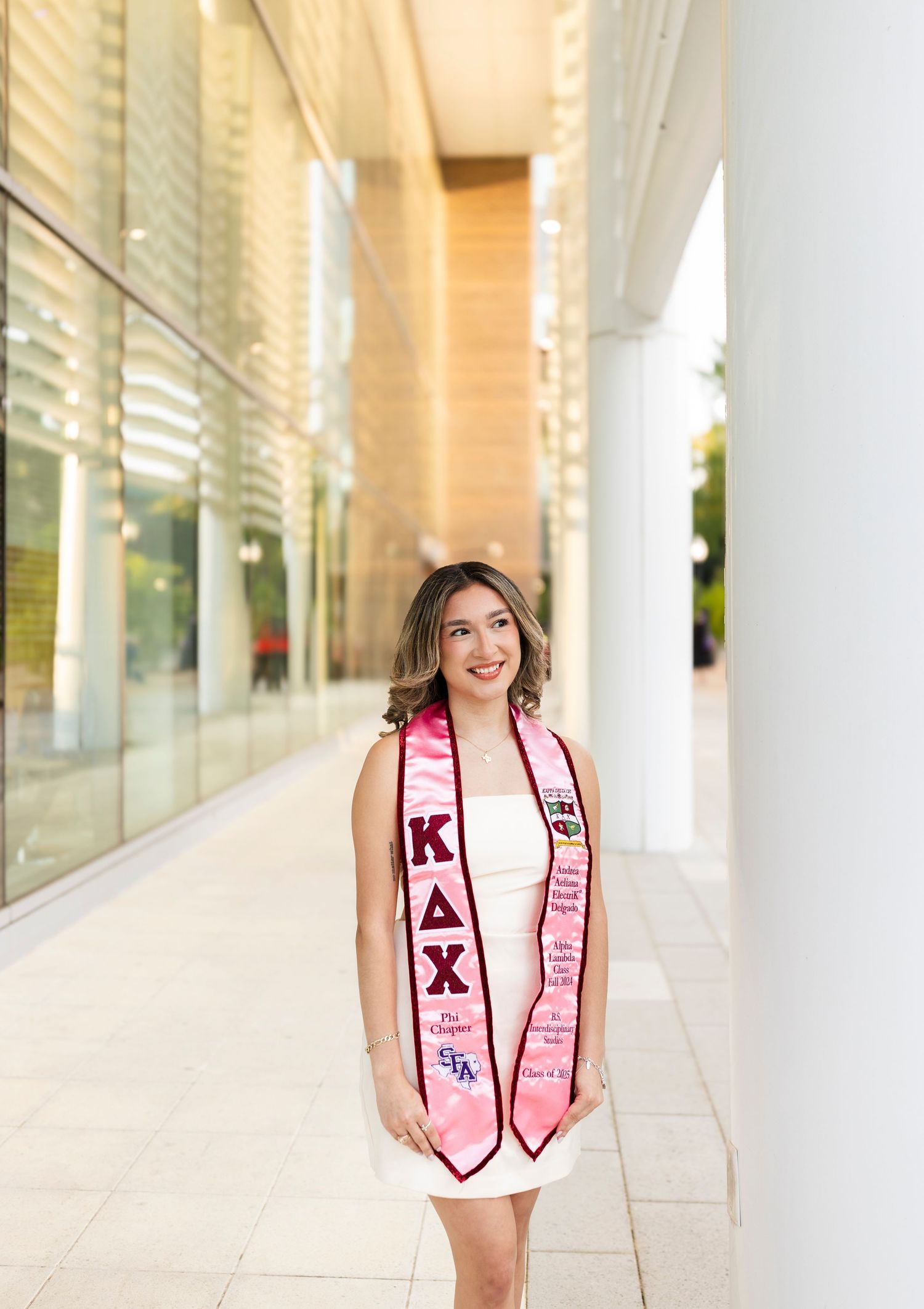 Smiling graduate wearing pink Kappa Delta Chi sorority stole poses in modern building corridor.