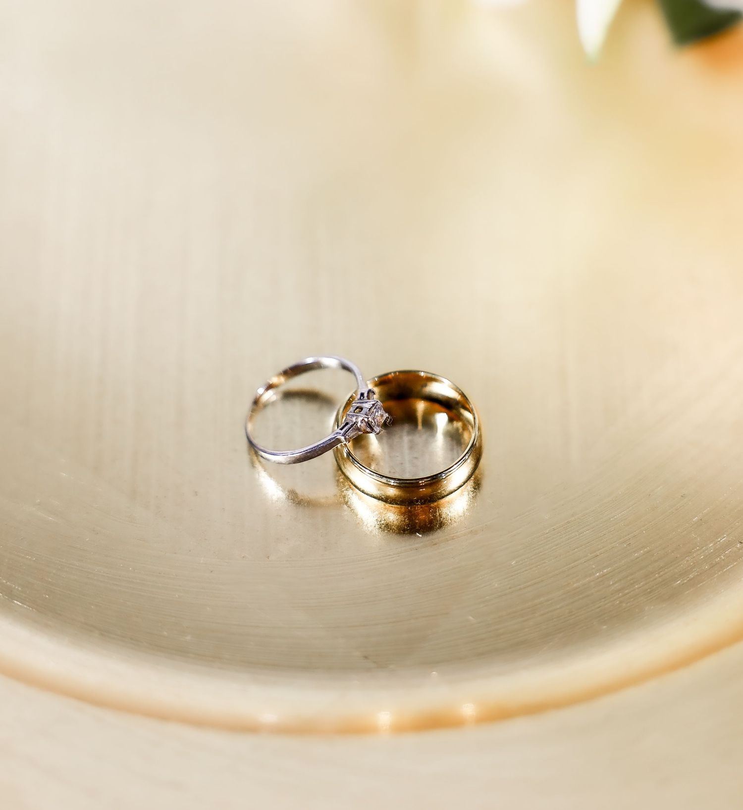 Two wedding rings, a gold band and a diamond engagement ring, rest together in a cream ceramic dish.