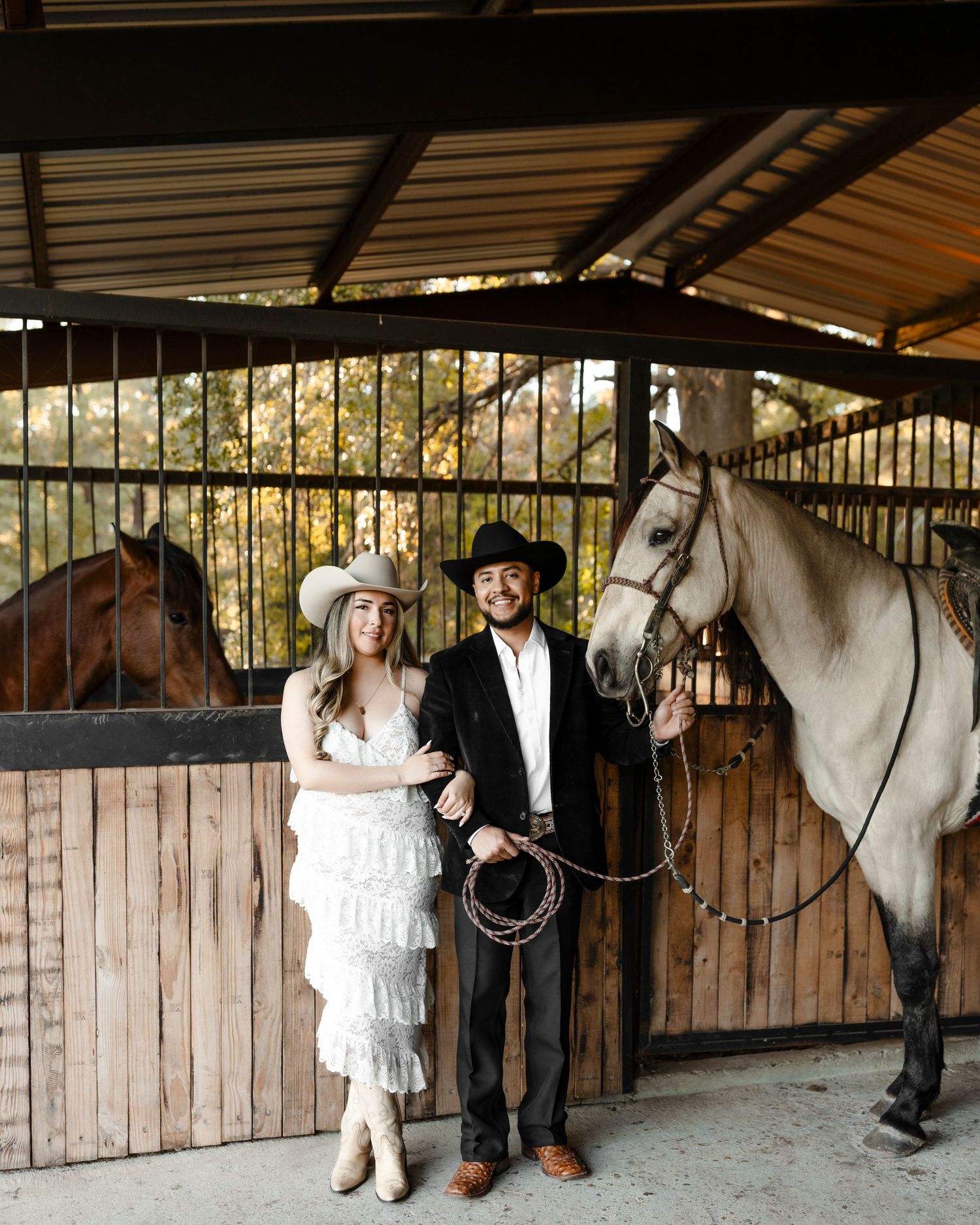 A couple in western attire pose with a horse inside a rustic stable, woman in white lace dress and cowboy hat.