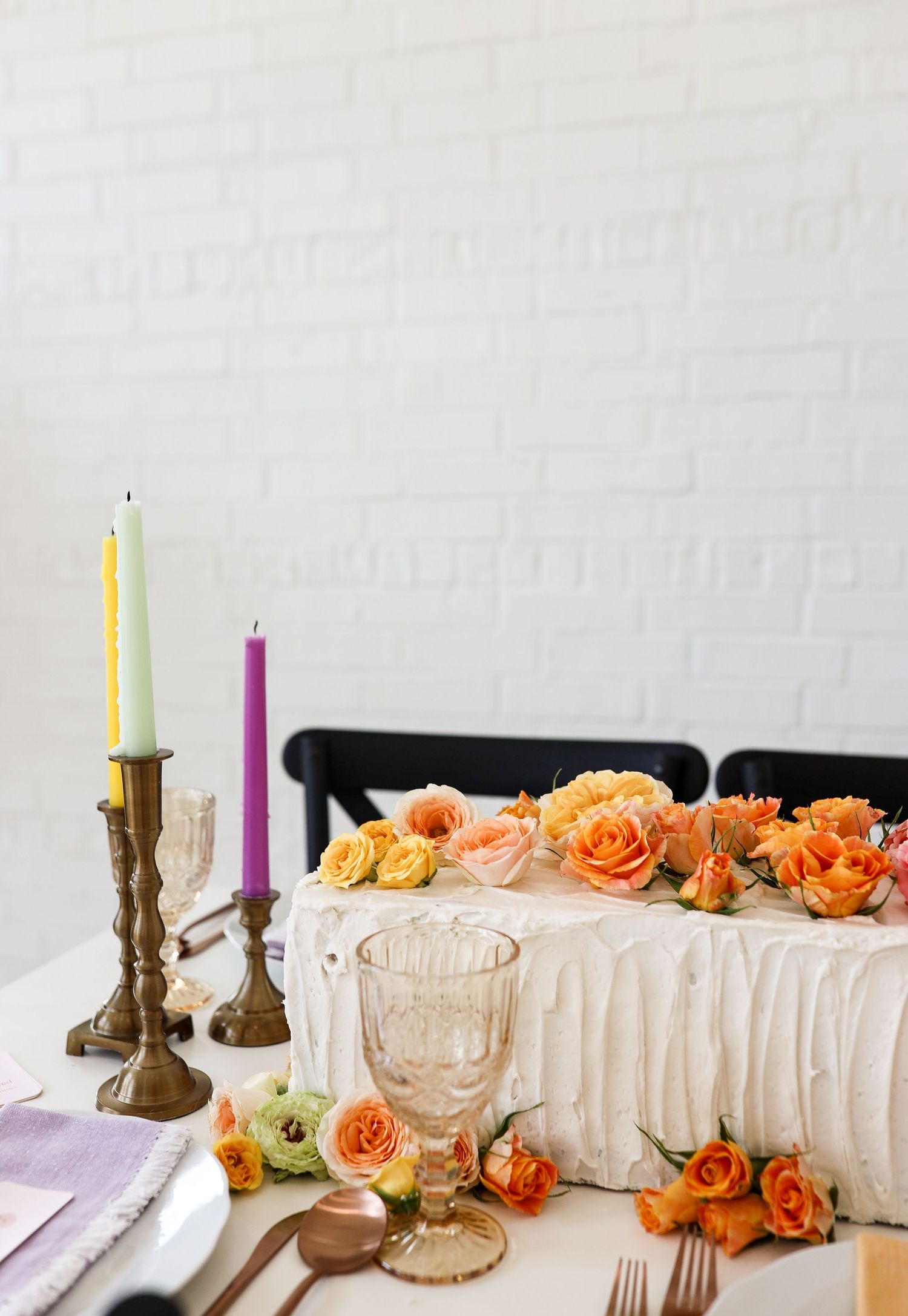 Wedding cake table with orange and peach roses, brass candlesticks with yellow and purple candles against white brick.