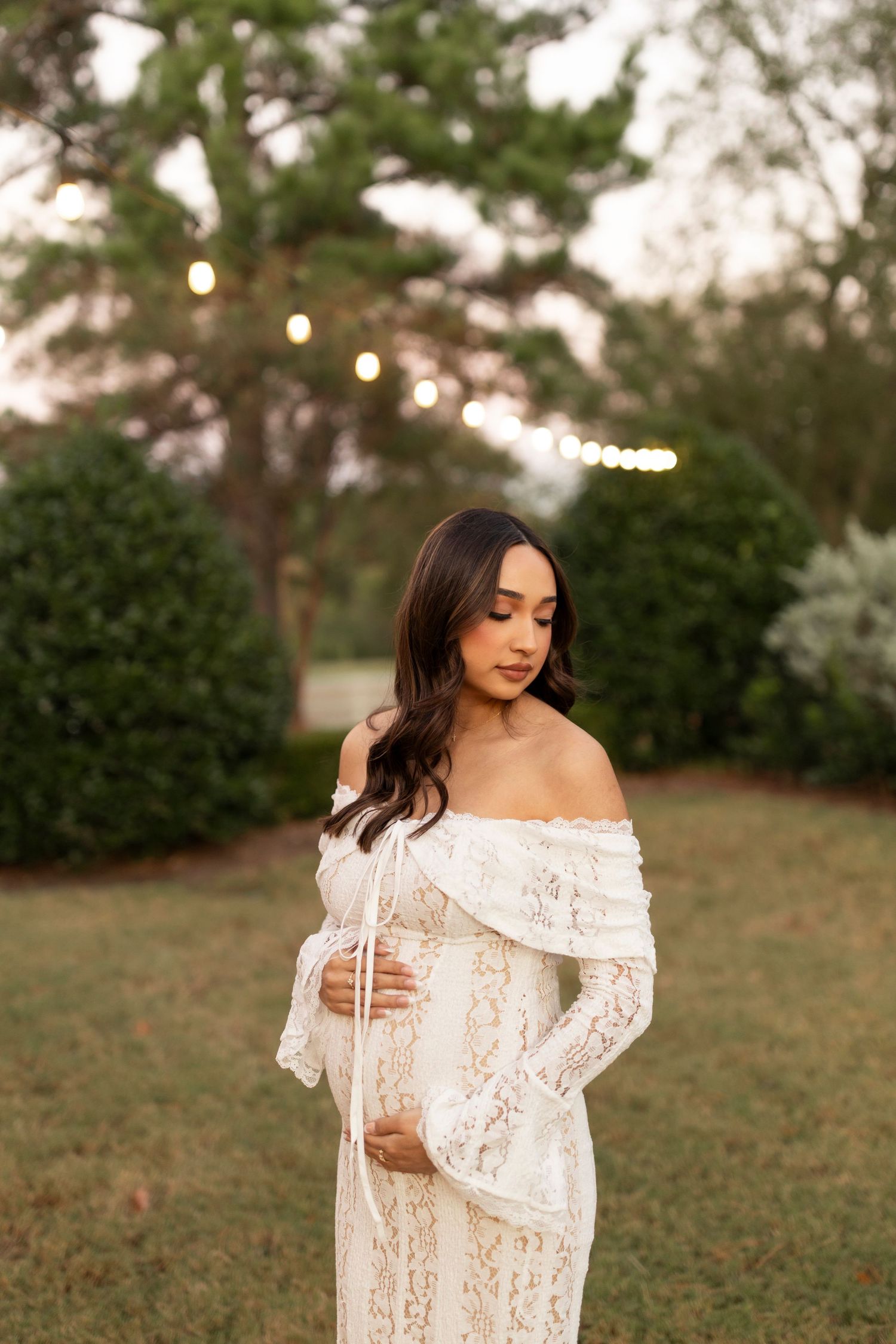 Pregnant woman in elegant off-shoulder white lace dress cradling her belly in a garden.