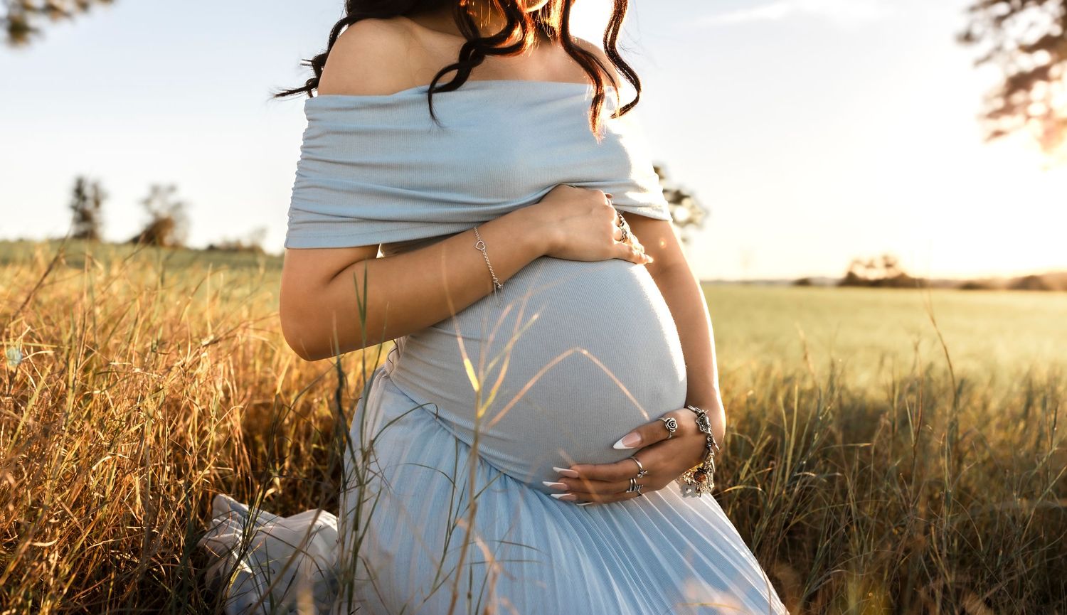 Pregnant woman in light blue off-shoulder gown cradling belly while sitting in golden wheat field at sunset.