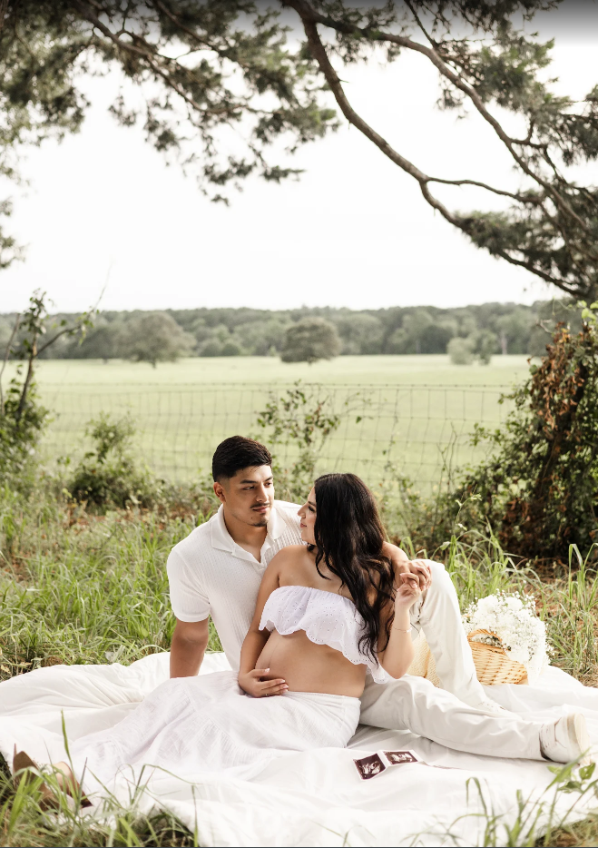 Expecting couple sitting on white blanket in scenic field during maternity photo session.