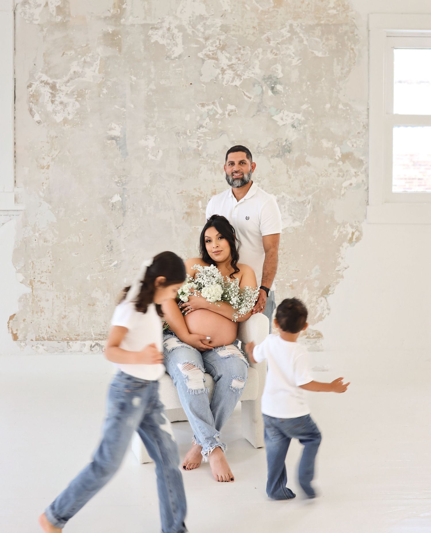 Family maternity photo with pregnant mother seated, father standing, children playing in white studio.