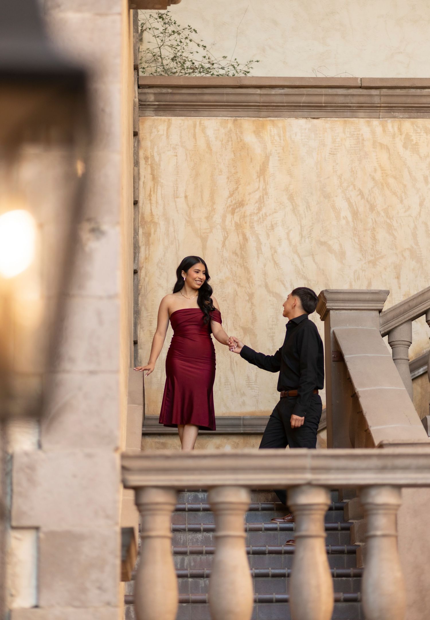 Couple holding hands on ornate stone staircase, woman in red strapless gown, man in black outfit.