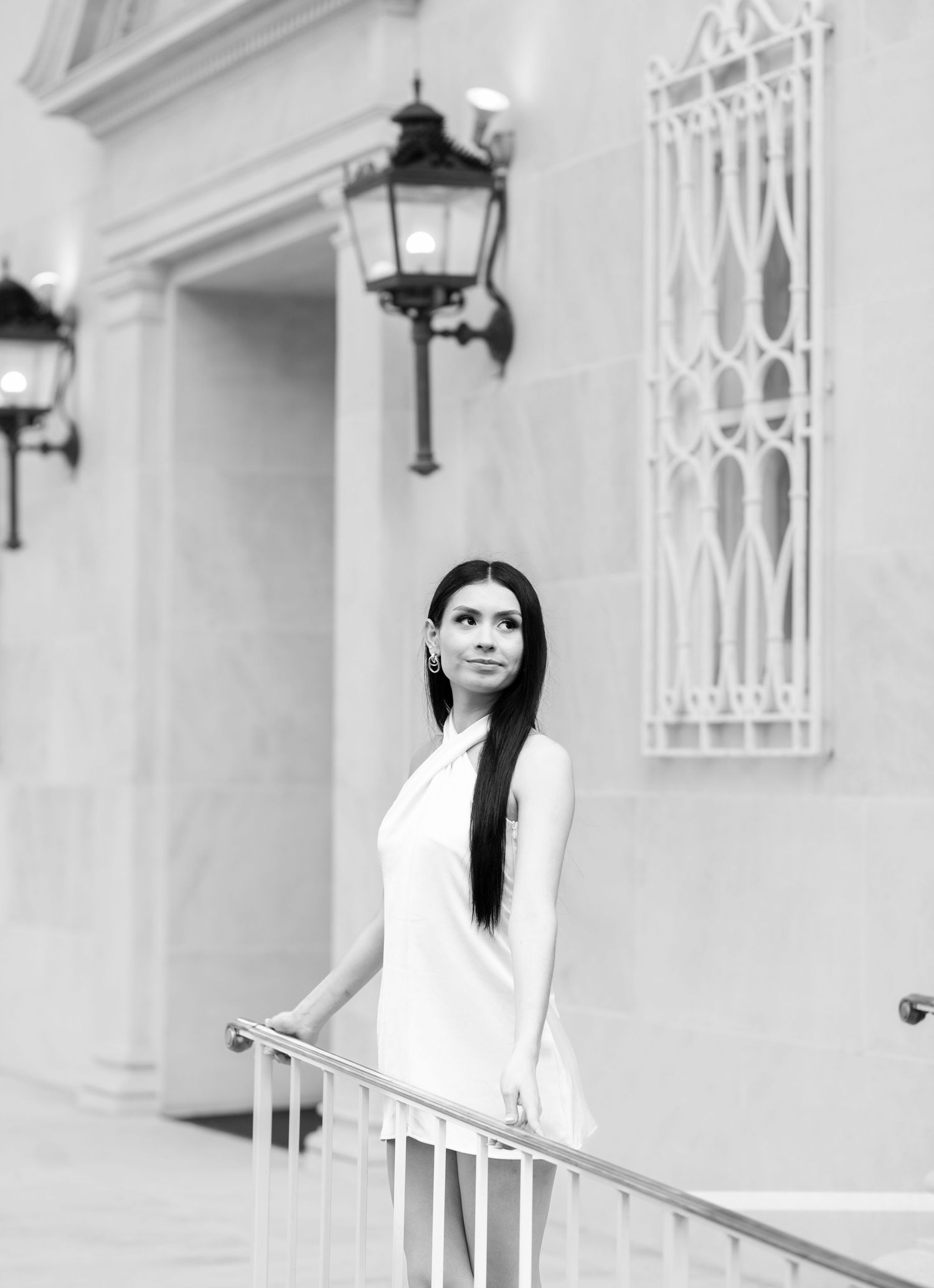 Black and white portrait of young woman in white dress standing by railing near ornate building.