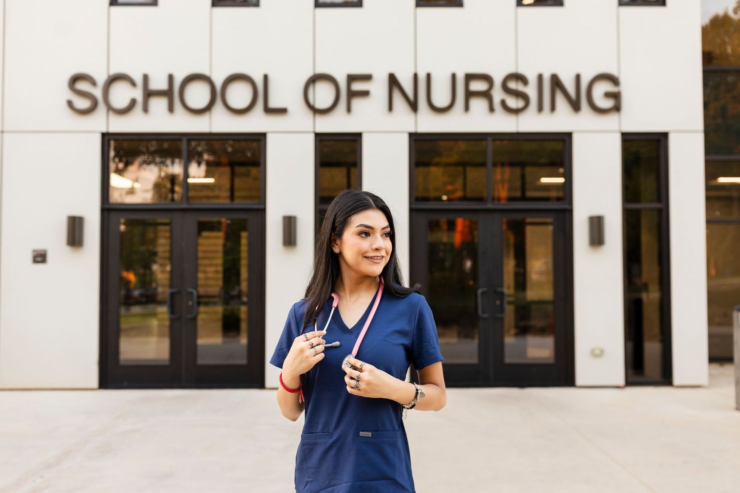Female nursing student in blue scrubs standing outside a School of Nursing building.