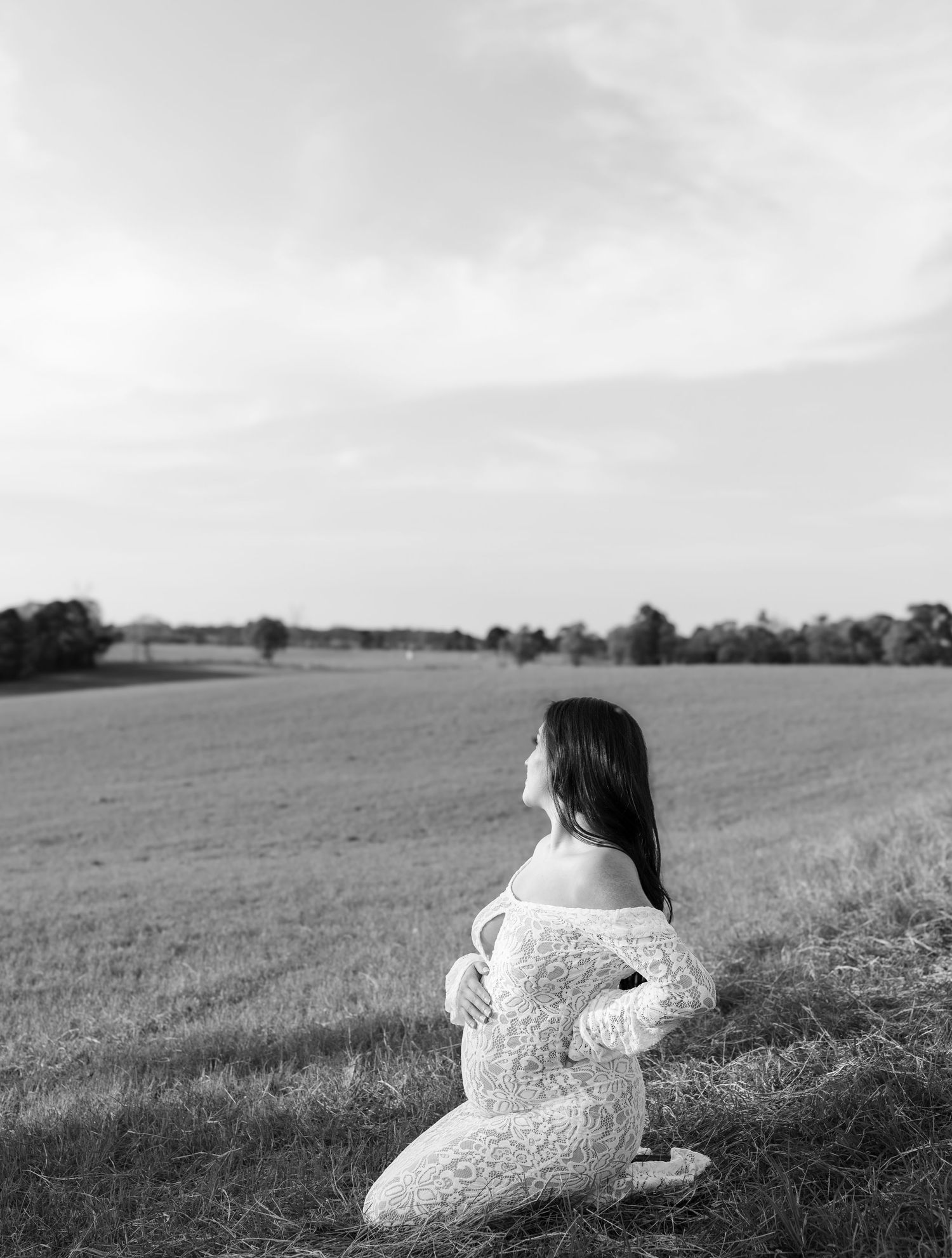 Black and white maternity photo of pregnant woman kneeling in lace bodysuit in open field.